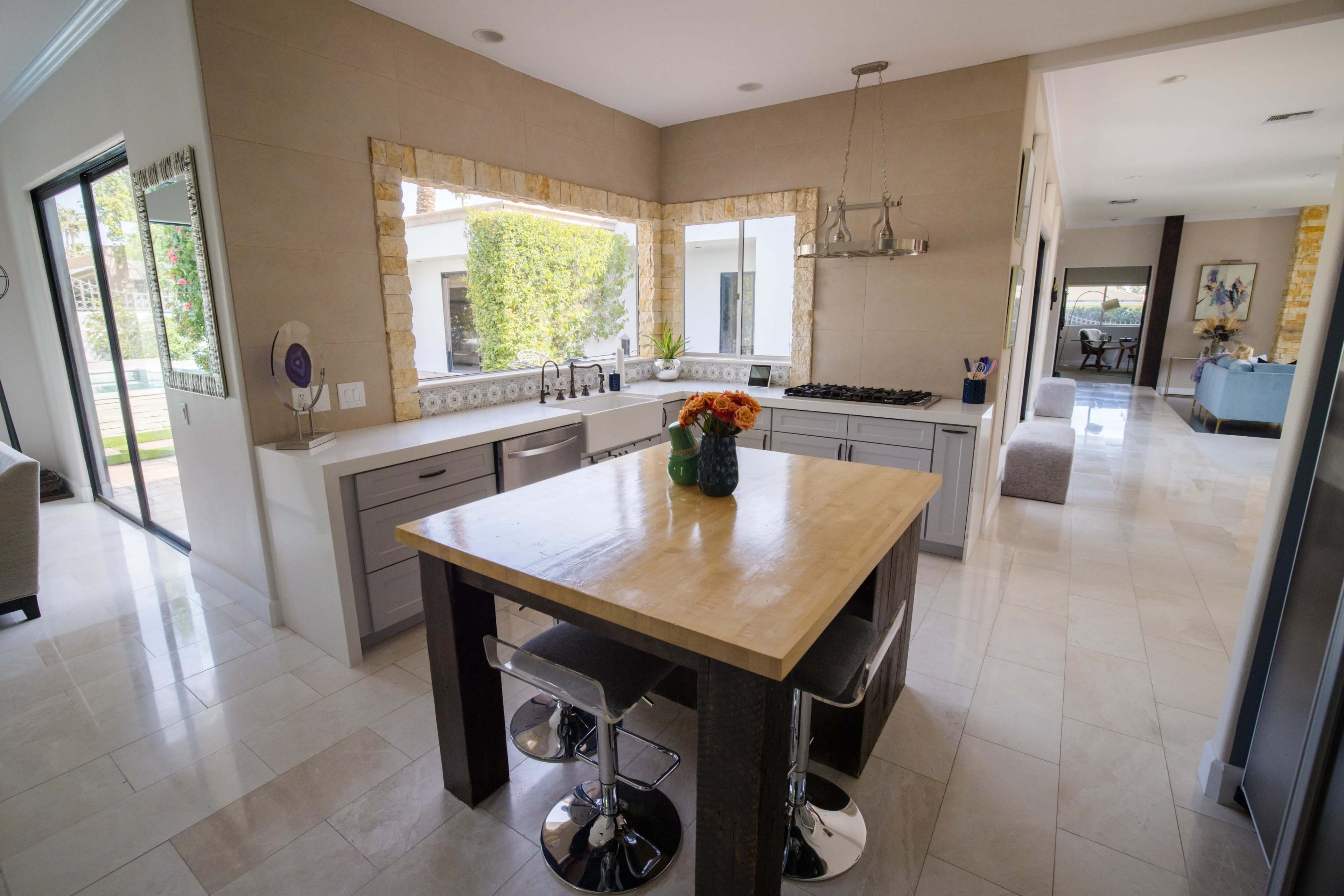 A modern kitchen features a central island with a light wooden surface and bar stools, surrounded by gray cabinets and large windows that provide a view of a patio area.
