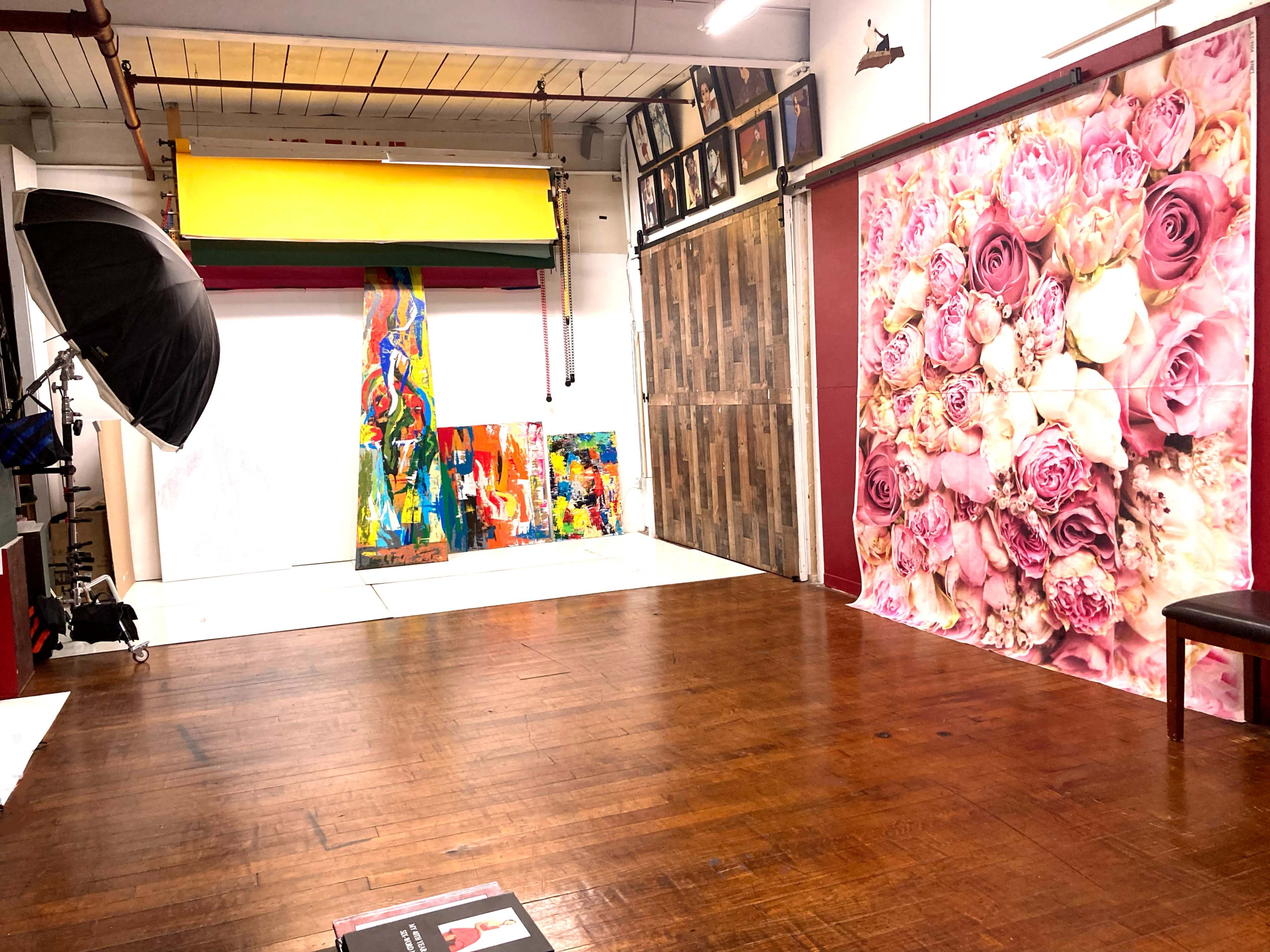 A photography studio featuring a wooden floor, a backdrop with pink roses, and multiple vibrant paintings set against a white backdrop.