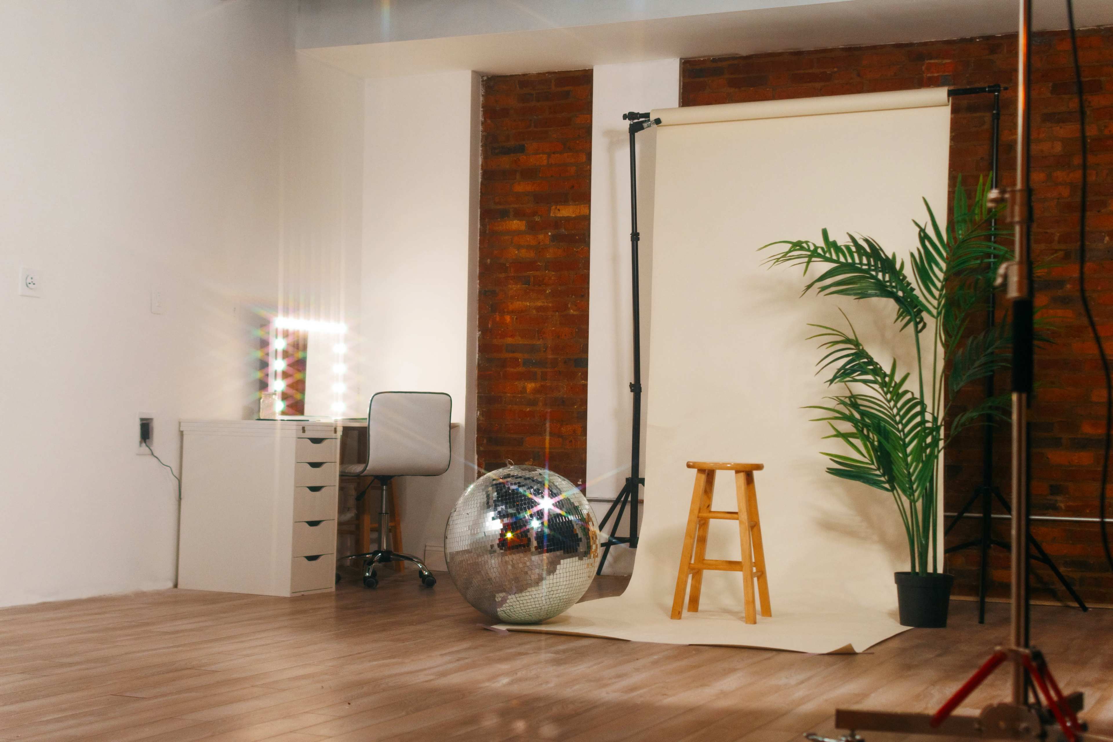 The scene features a minimalistic studio setup with a stool, a mirrored disco ball, a plant, and a vanity table against a backdrop of beige and exposed brick walls.