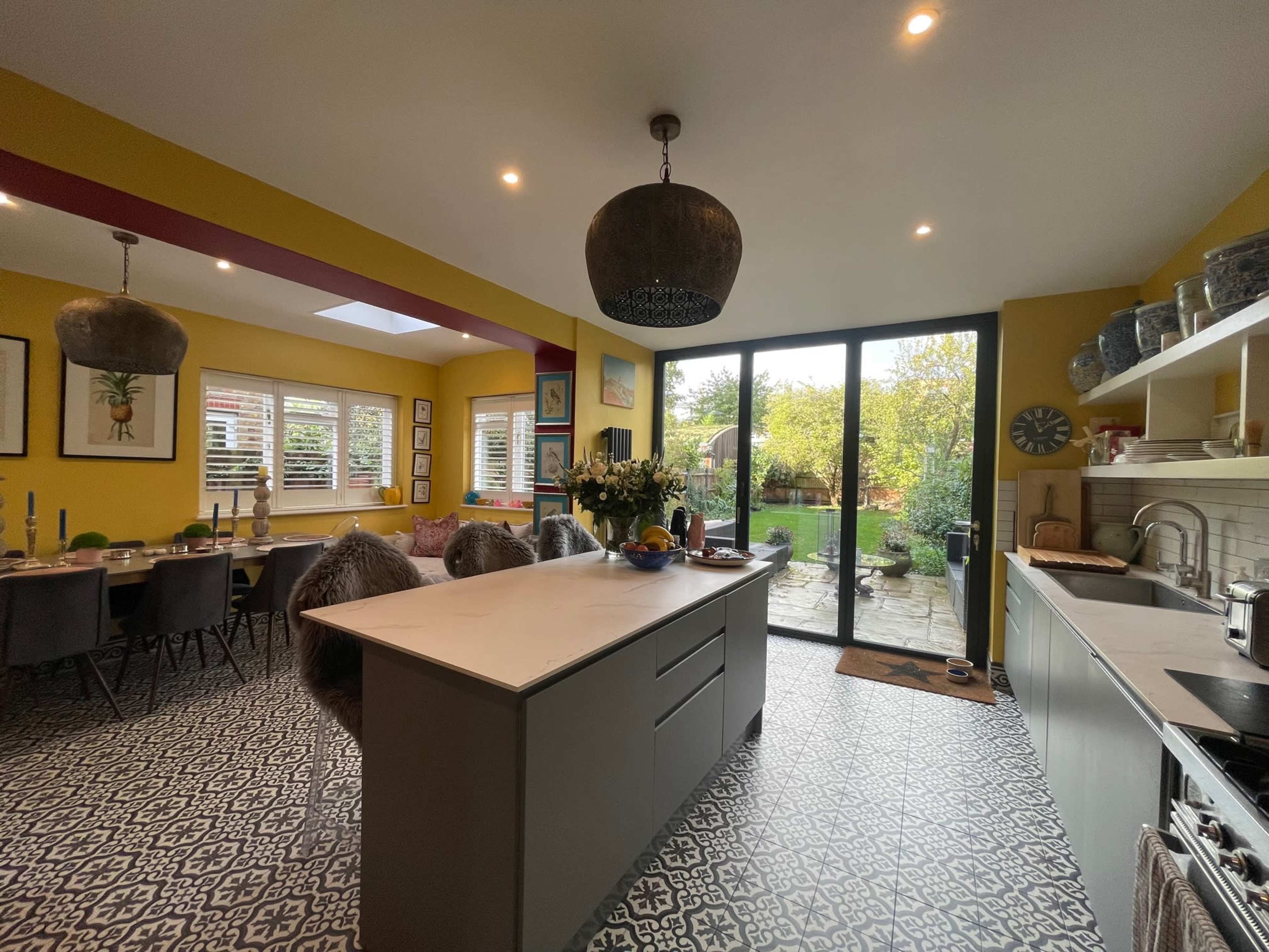 A modern kitchen features a central island with a dark countertop, dining table, and large glass doors that open to a garden.