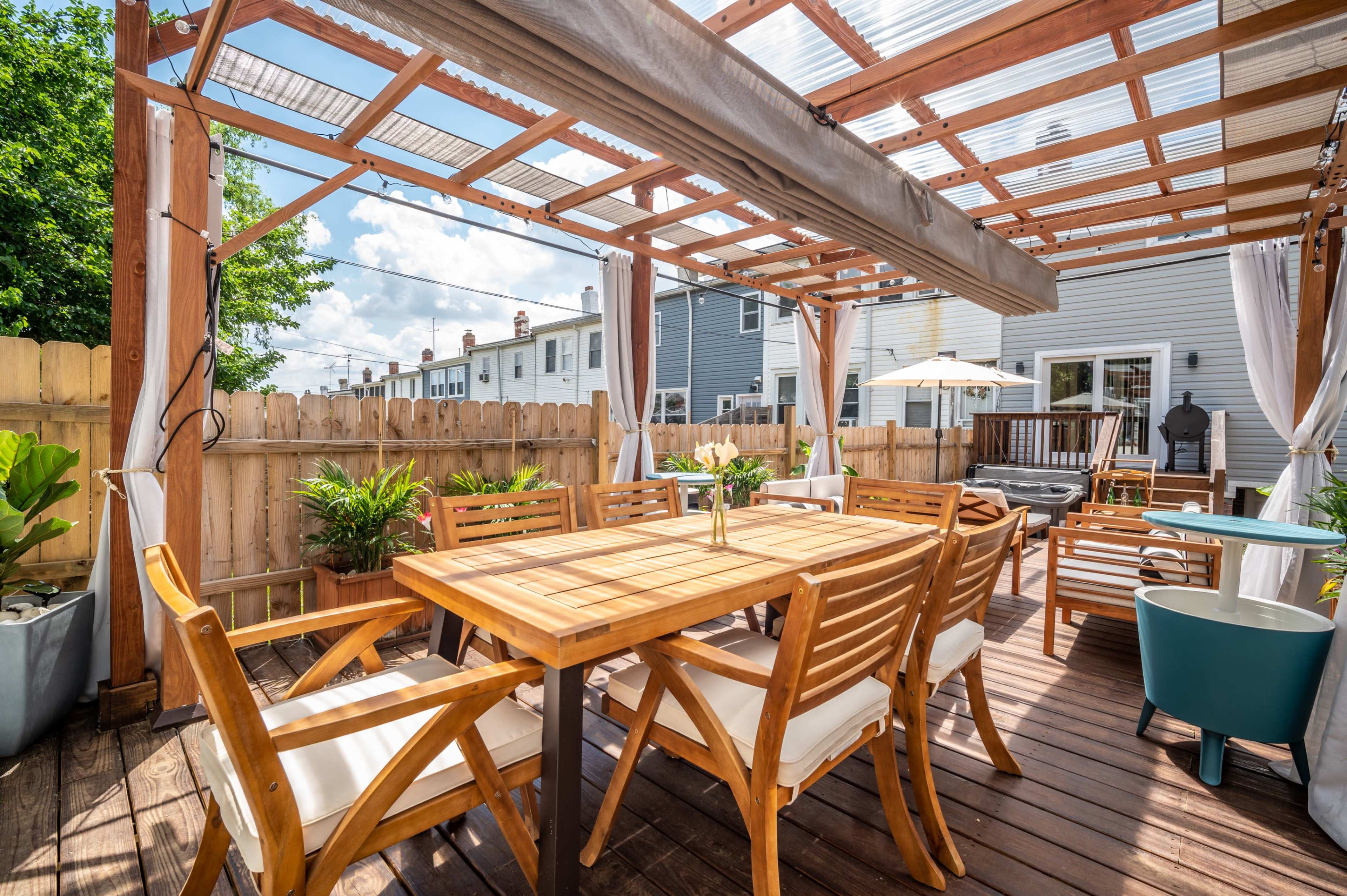 A wooden outdoor patio with a table set for dining, surrounded by plants and covered by a pergola.