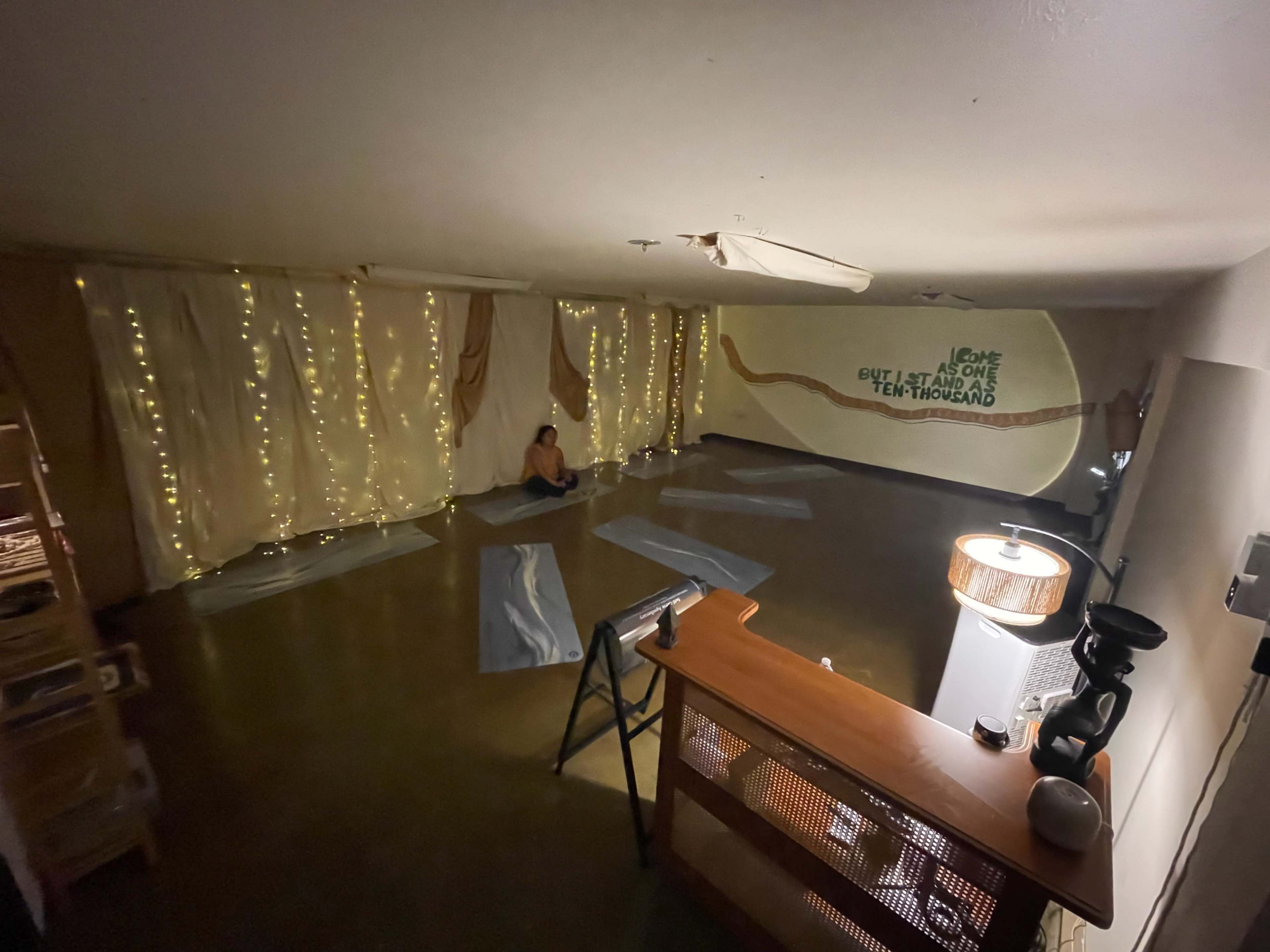 A yoga studio with dim lighting features yoga mats on the floor and a person sitting in meditation against softly draped fabric and string lights.