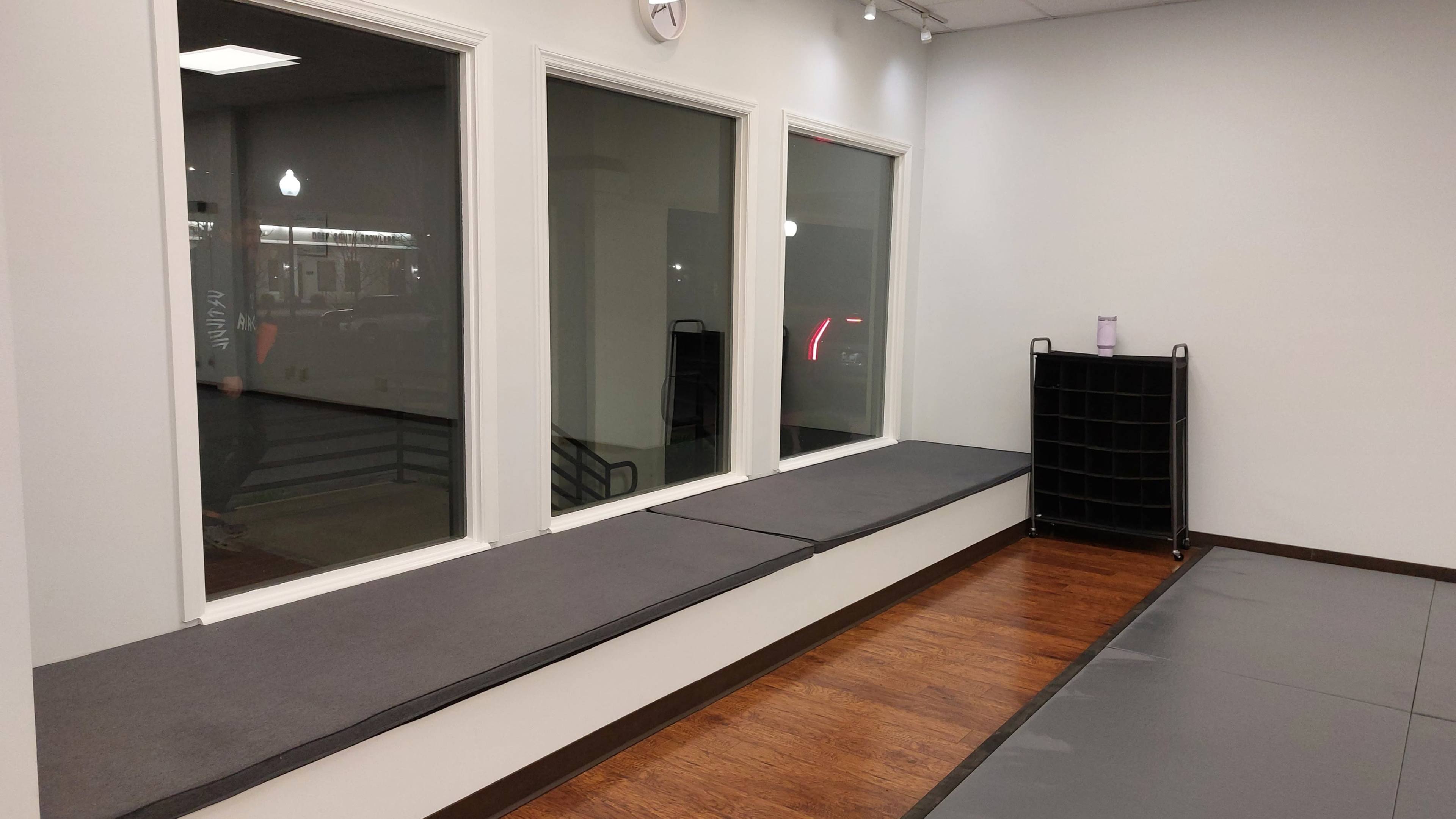 The image shows a minimalist room with large windows on one wall, a low ledge with a cushion, and a shelf in the corner.