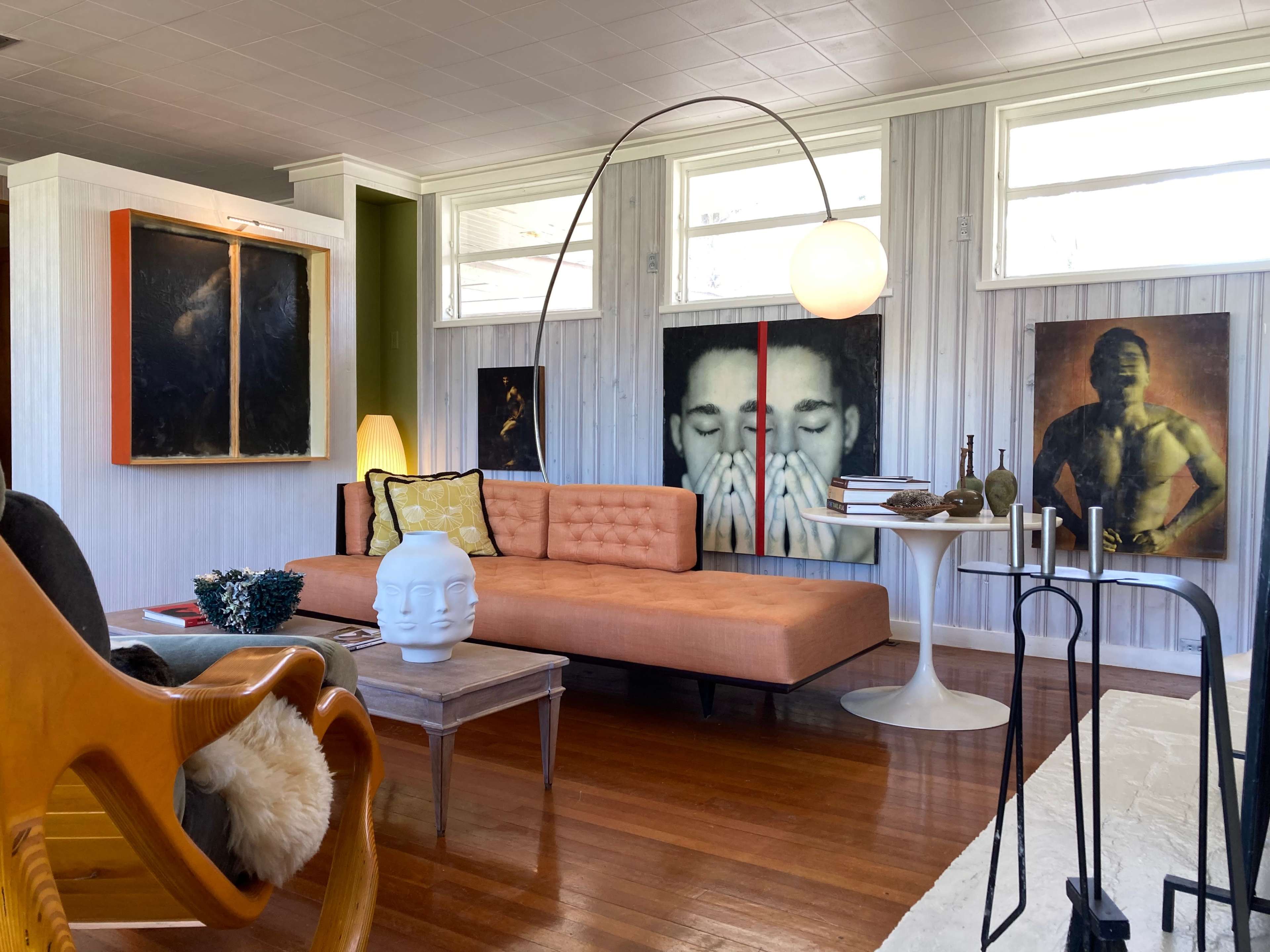 A mid-century modern living room features a patterned sofa, a round dining table, and several large framed portraits on the walls.