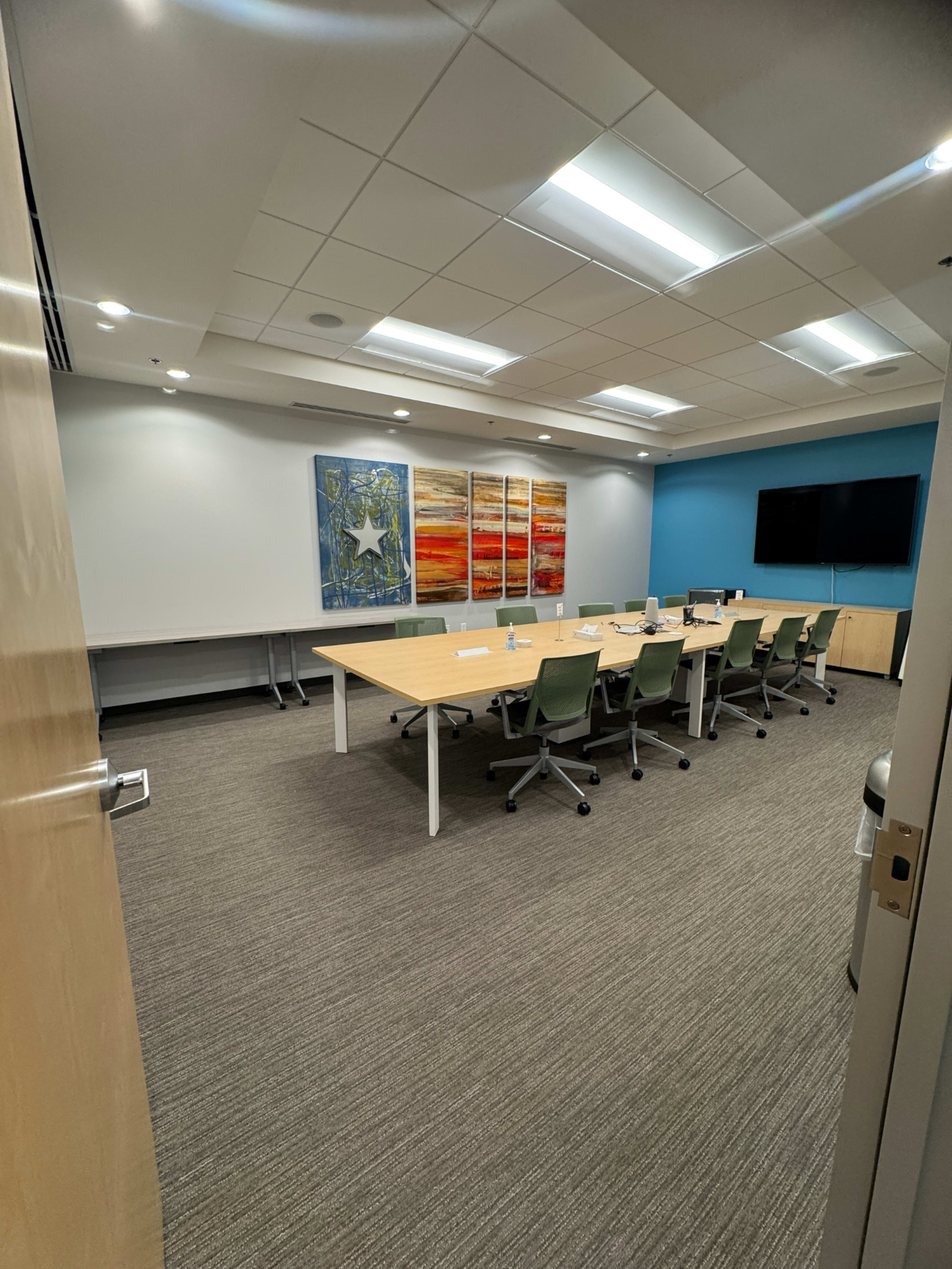 A conference room features a long table surrounded by rolling chairs, with three large paintings on the wall and a TV mounted in the corner.