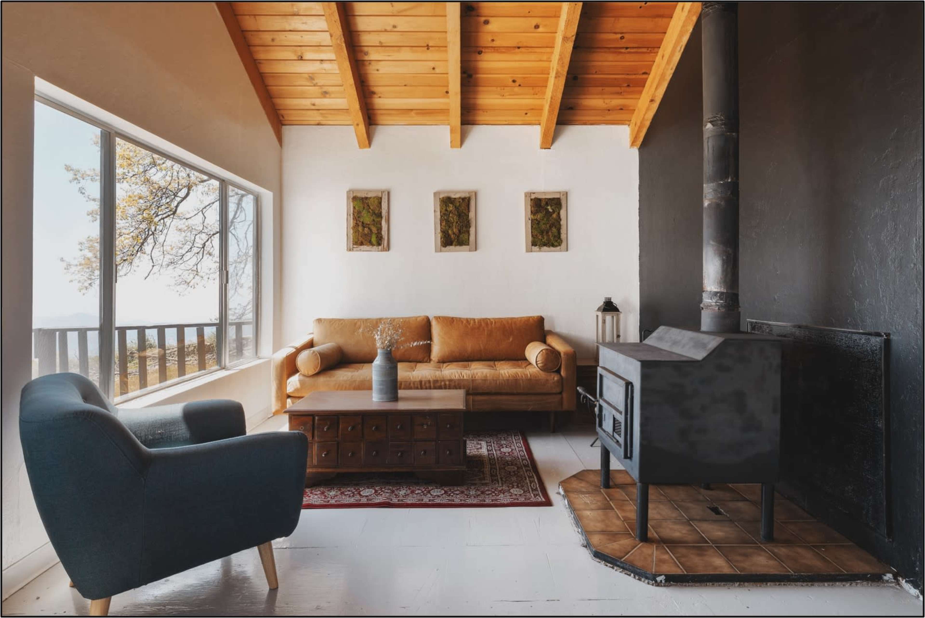 A cozy living room featuring a brown sofa, a blue armchair, a wooden coffee table, and a black wood stove, with large windows allowing natural light to enter.
