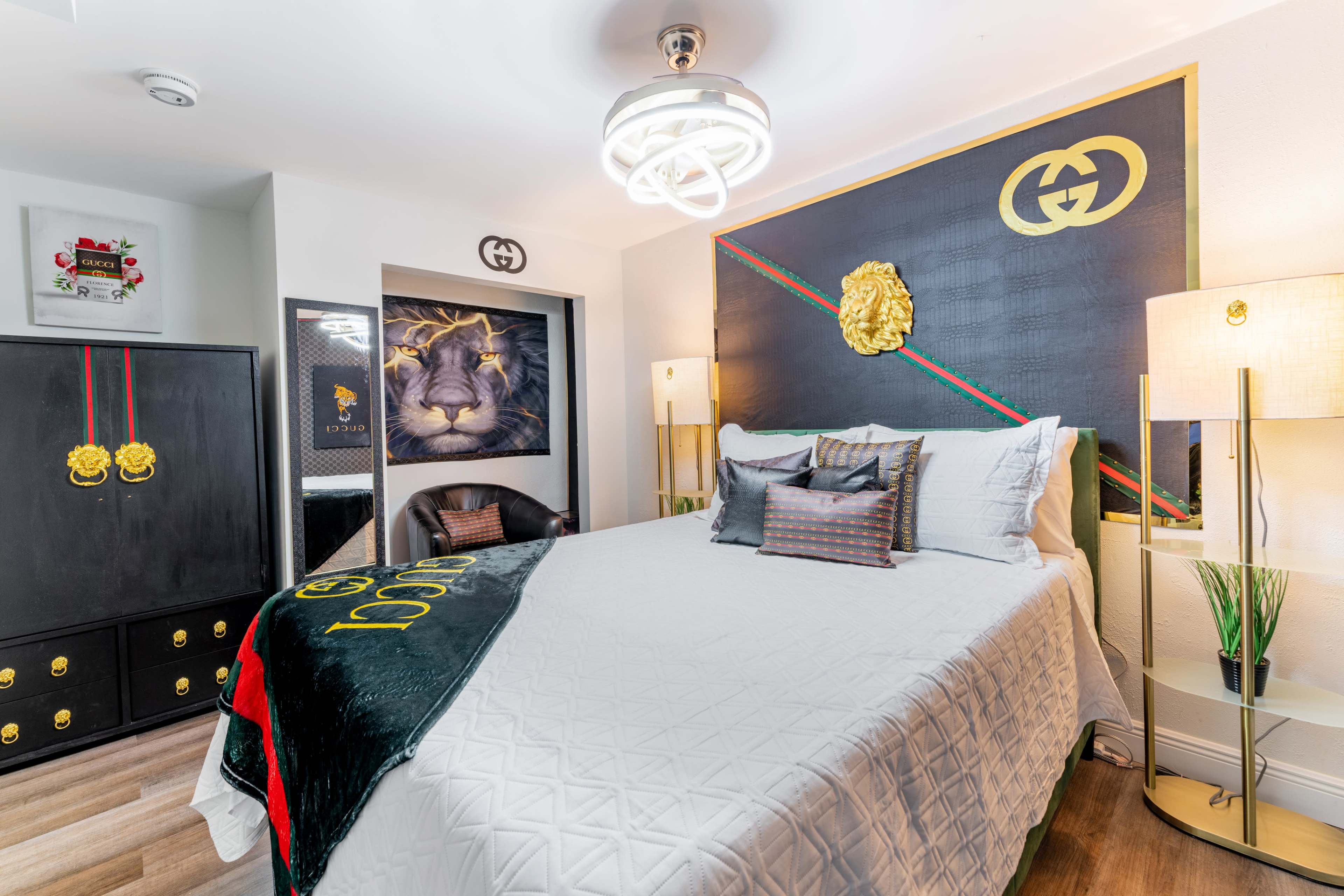 A bedroom featuring a large bed with white bedding, a black and gold dresser, and a decorative wall with a lion and the Gucci logo.
