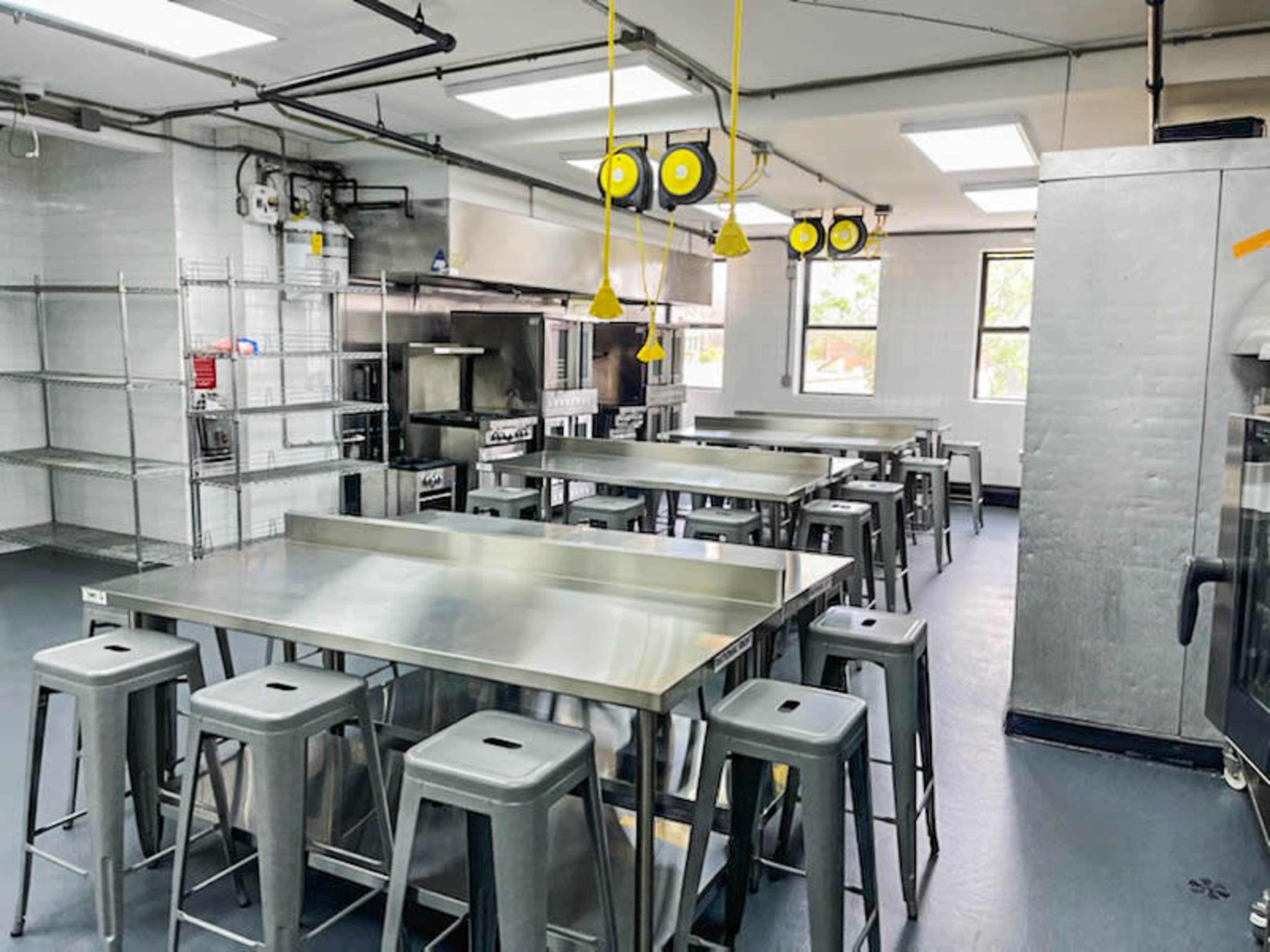 A spacious commercial kitchen equipped with stainless steel tables, metal stools, and industrial appliances.
