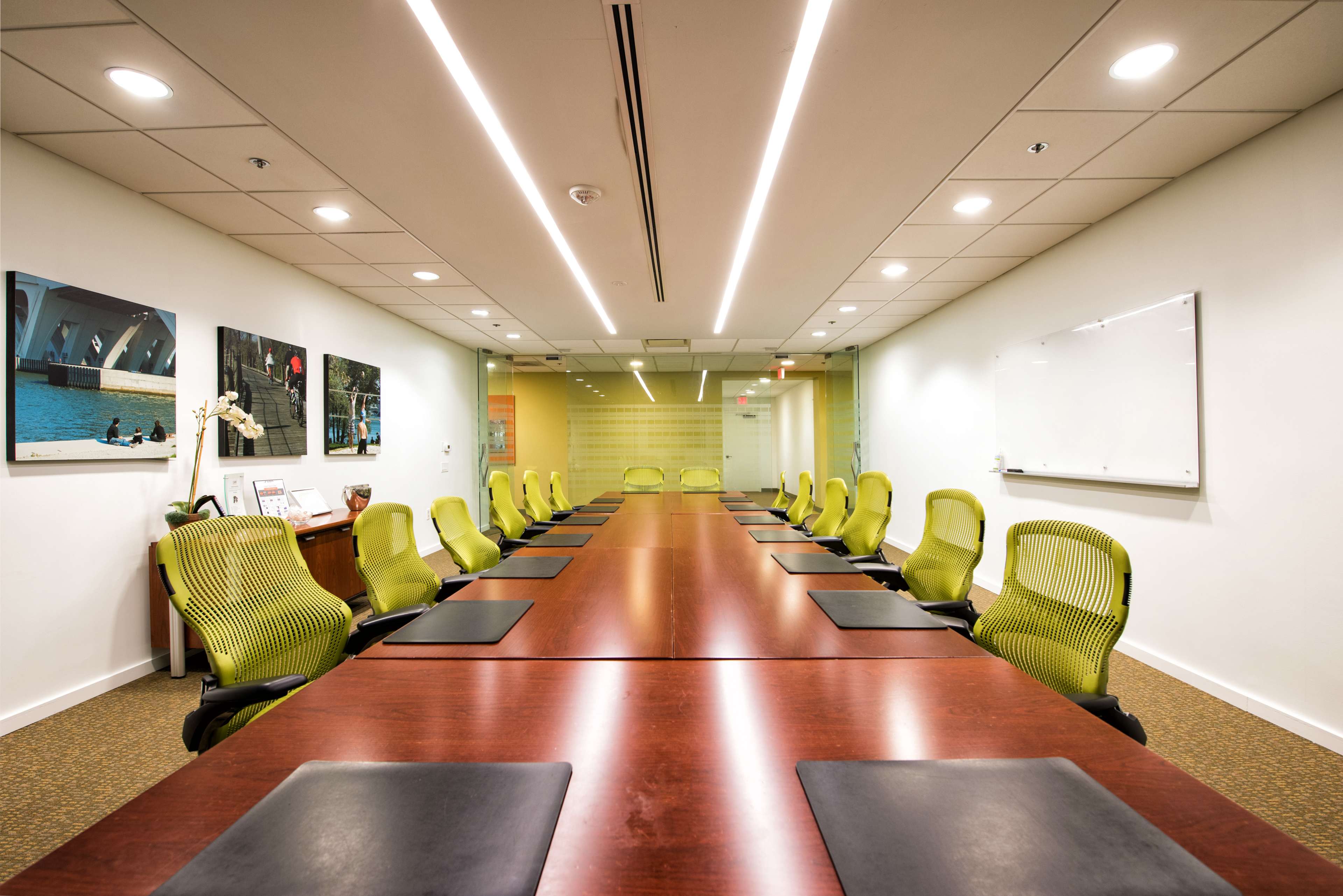 A long conference table with green chairs is arranged in a well-lit meeting room featuring artworks on the walls.