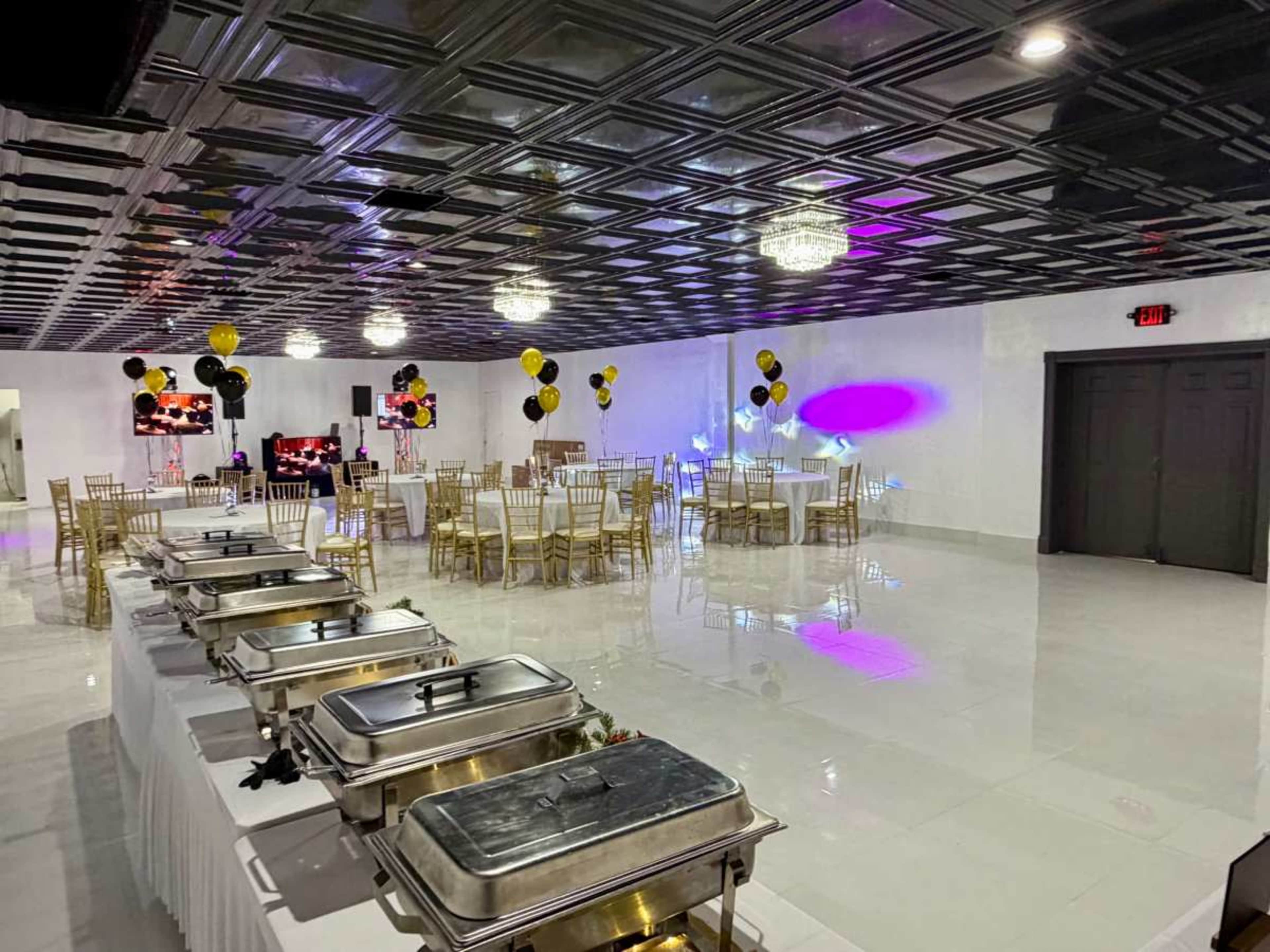 The image shows a spacious event hall set up for a celebration, featuring tables with white tablecloths, gold chairs, decorative balloons, and a buffet area with metal food warmers.