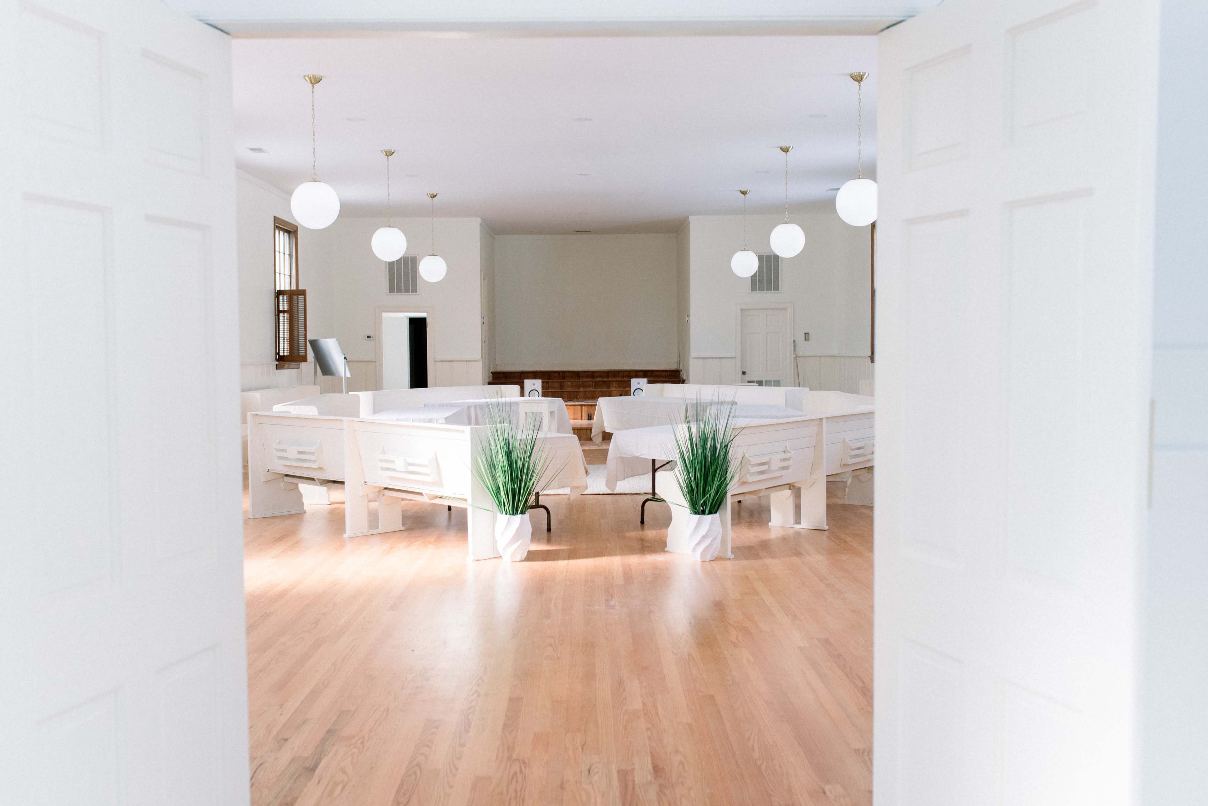An empty, bright room with white walls and wooden floors, featuring round lights hanging from the ceiling and tables arranged in a circular layout.