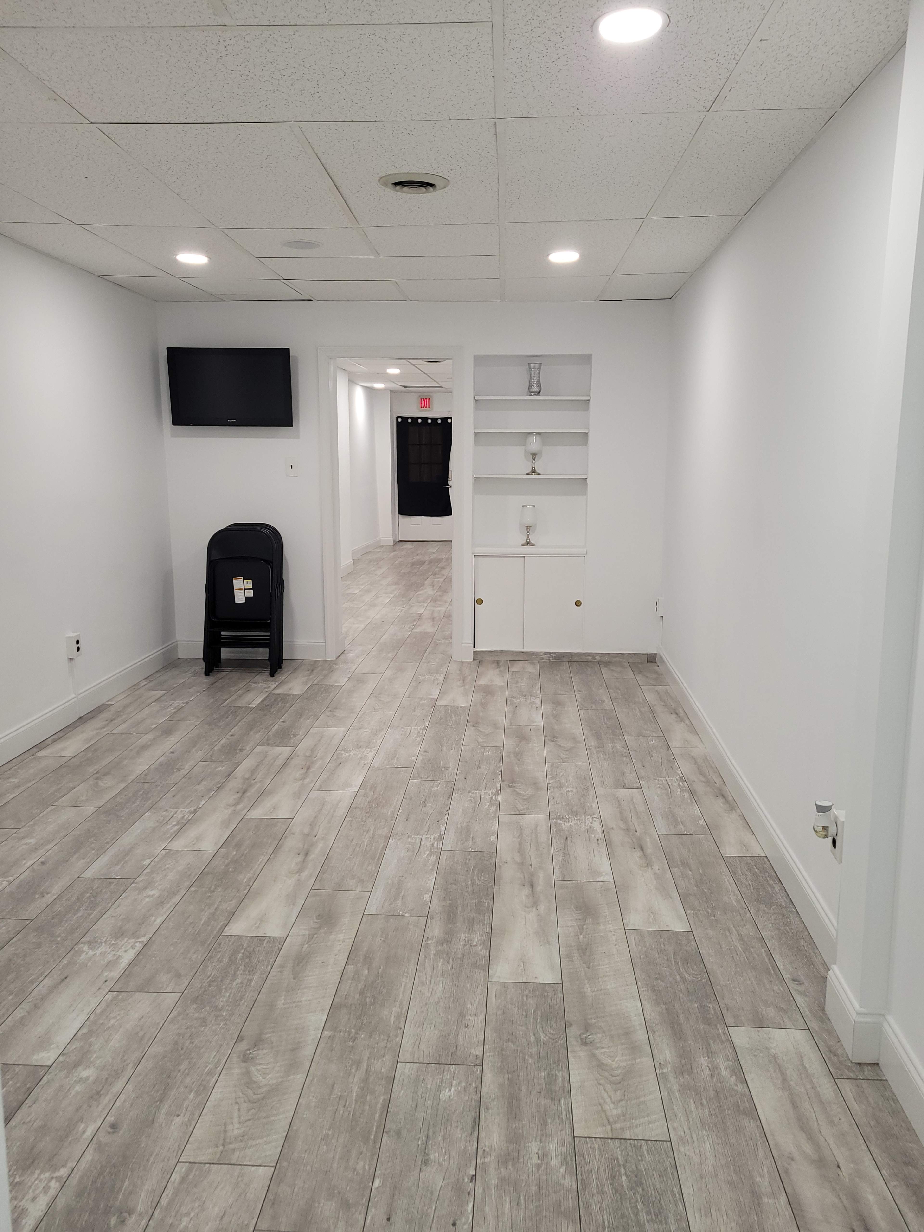The image shows a clean, modern hallway with light-colored walls, wood-like flooring, a television mounted on the wall, and a built-in shelving unit.