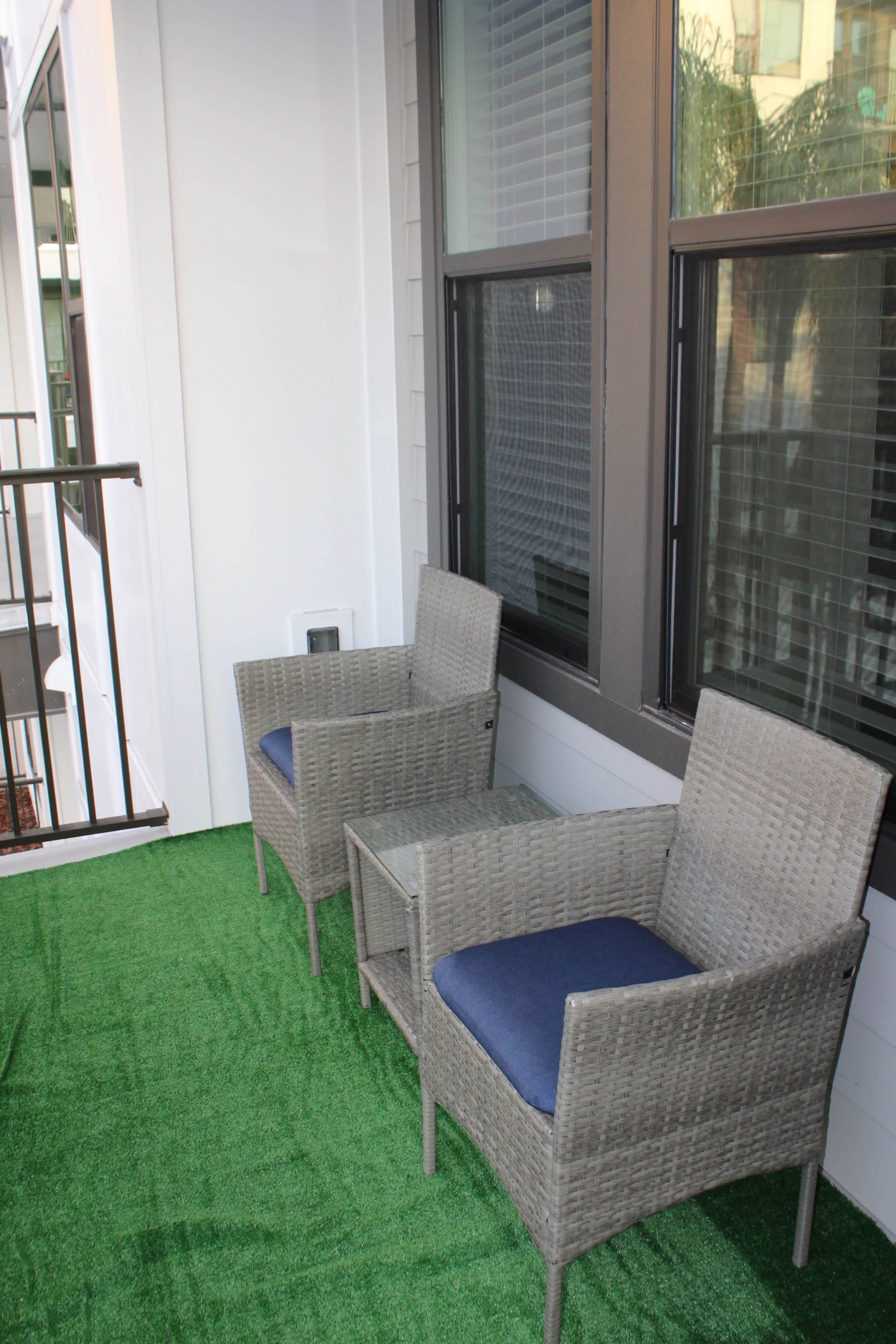 A small balcony with two wicker chairs and a table, surrounded by green synthetic grass.