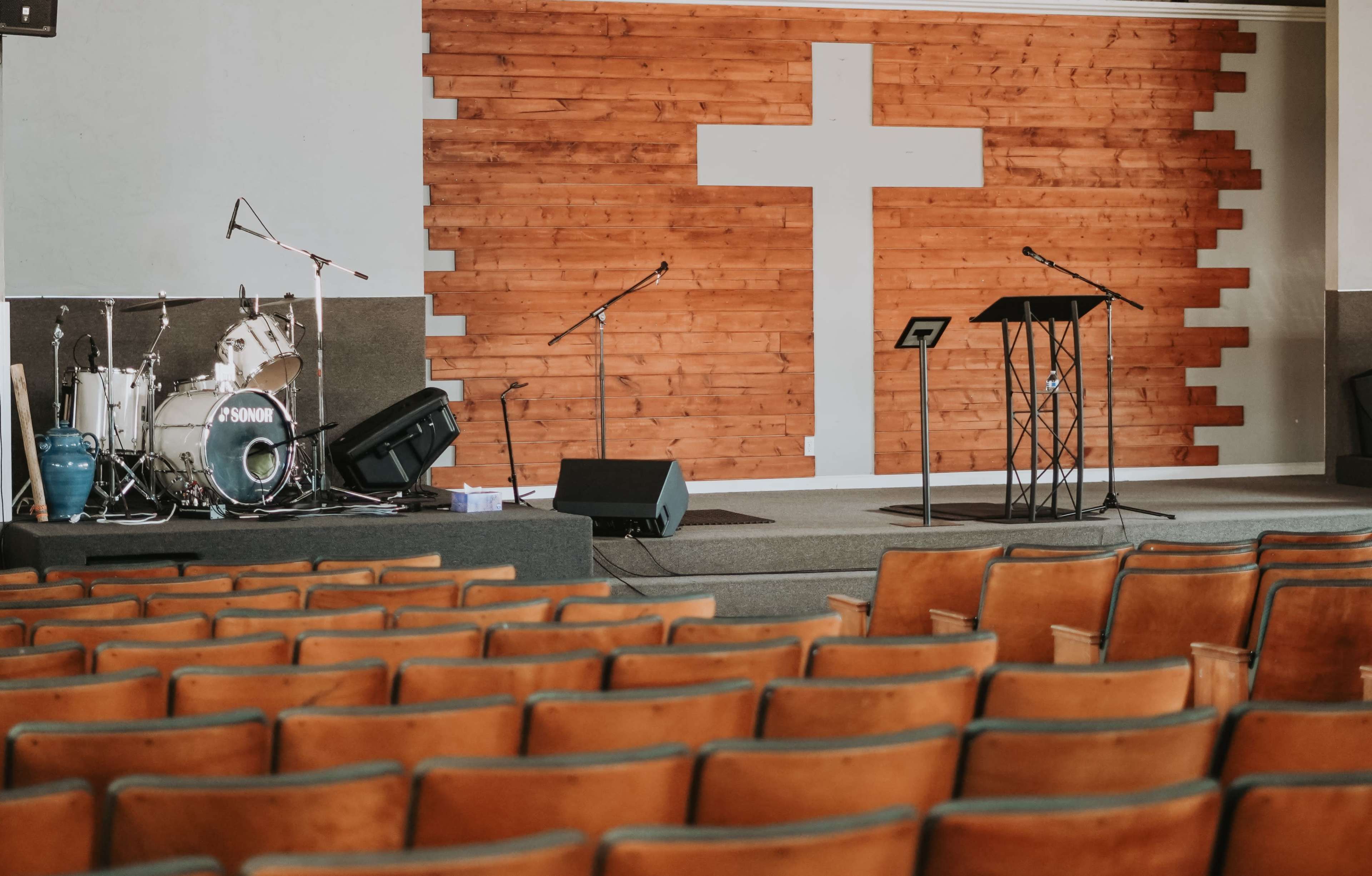 A church interior with a wooden cross on the wall, a stage set for a service featuring a drum set, microphones, and a podium.