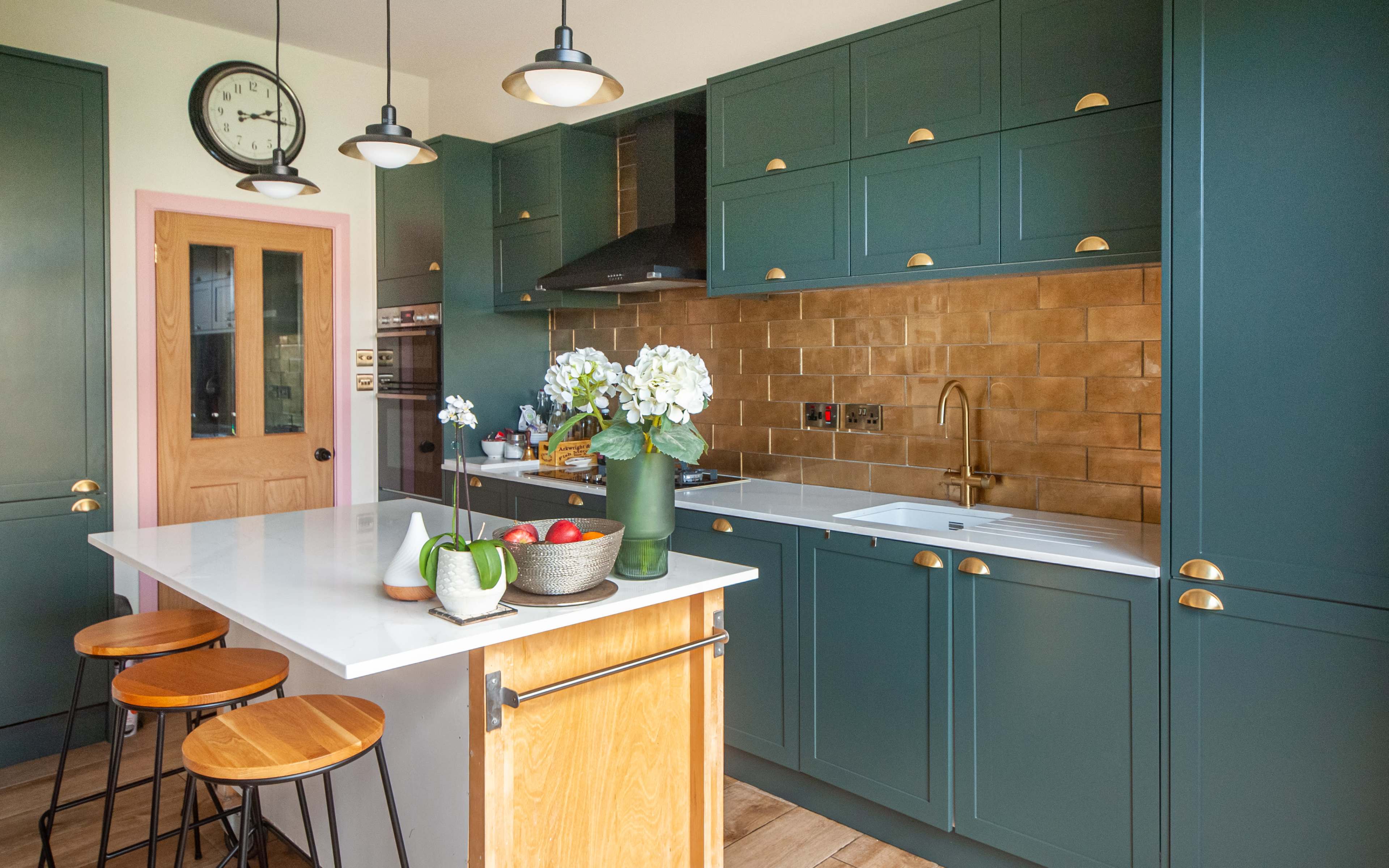 The image shows a modern kitchen featuring green cabinets, a white countertop with bar stools, and a gold-accented backsplash.