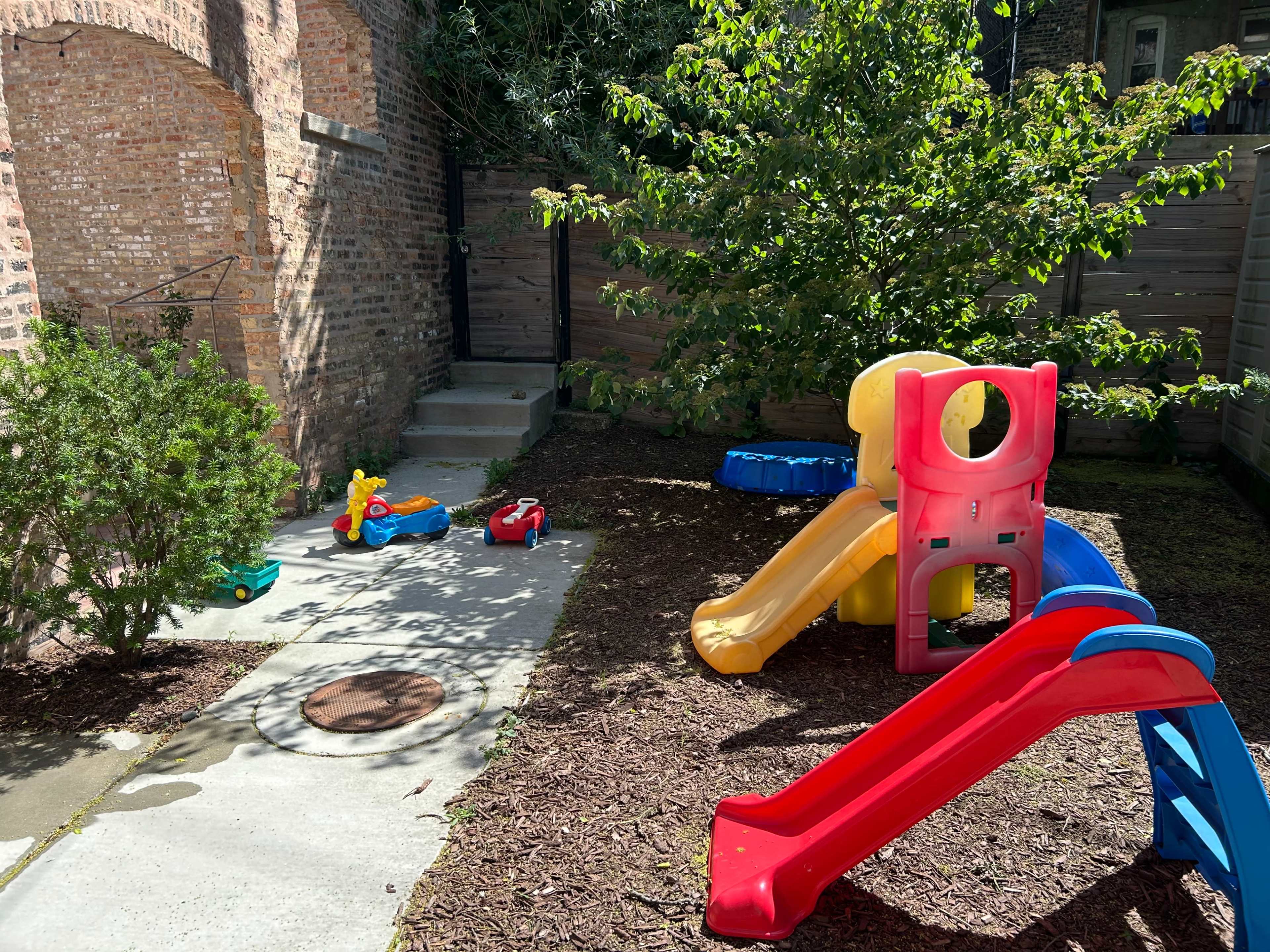 A small backyard features colorful children's play equipment, including a slide, a ride-on toy, and a small playhouse surrounded by mulch and greenery.