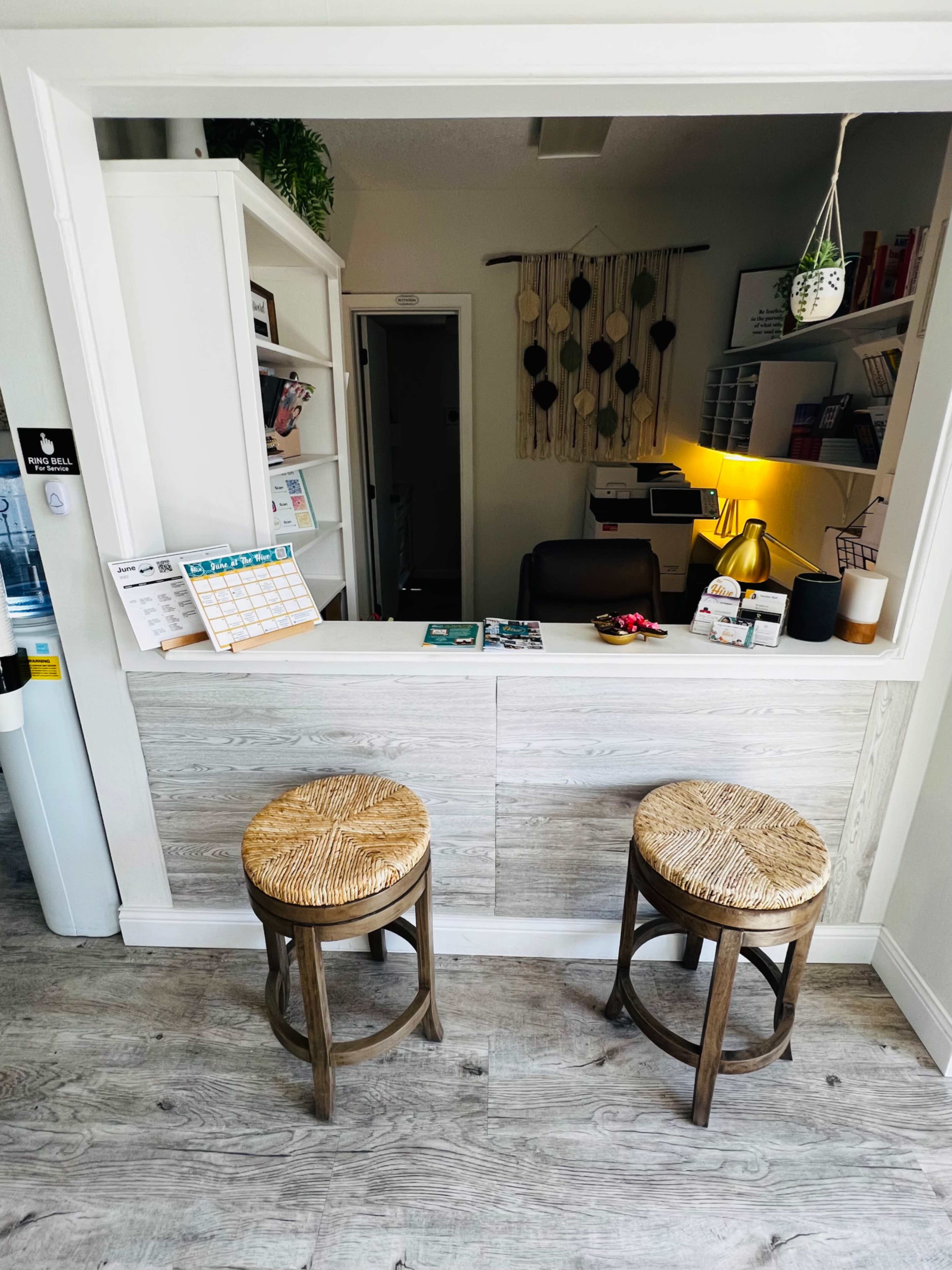 The image shows a small reception area featuring two woven stools and a counter with a calendar and paperwork on it.