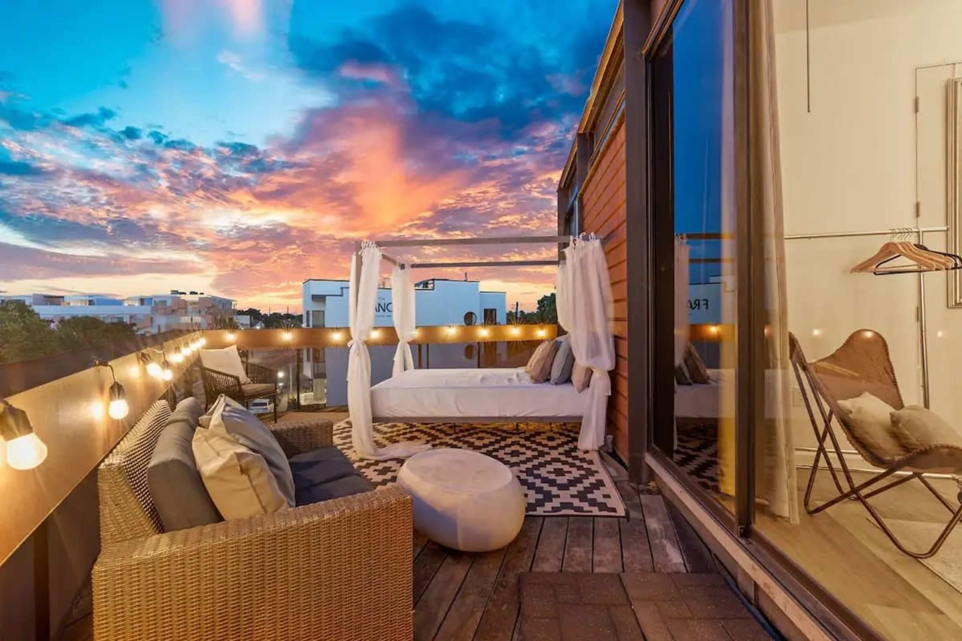 A balcony features a seating area with woven furniture, a lounge chair, and decorative lights, all set against a colorful sunset sky.