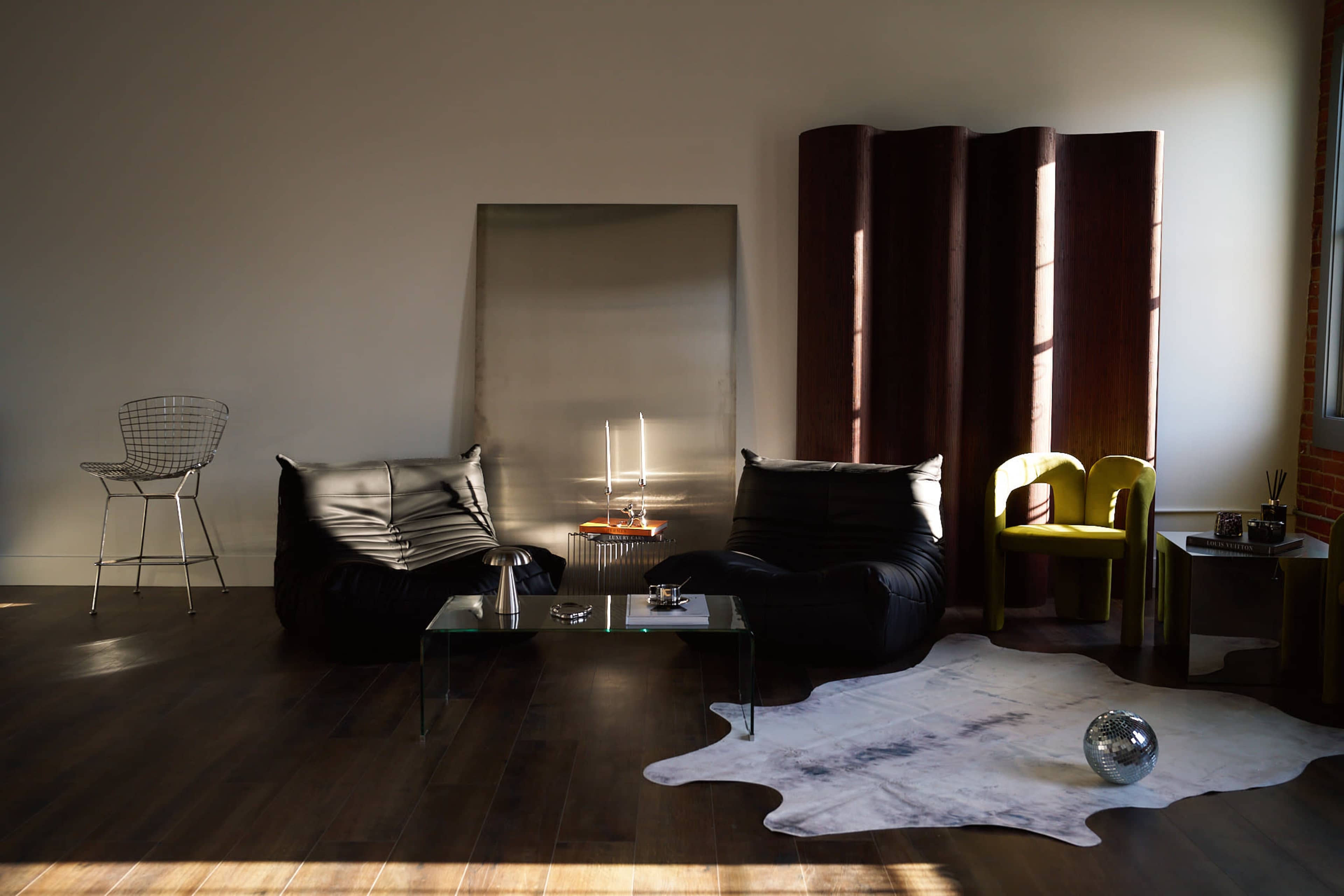 A modern living room features two black bean bag chairs, a glass coffee table with decorative items, and a cowhide rug, illuminated by sunlight filtering through large windows.