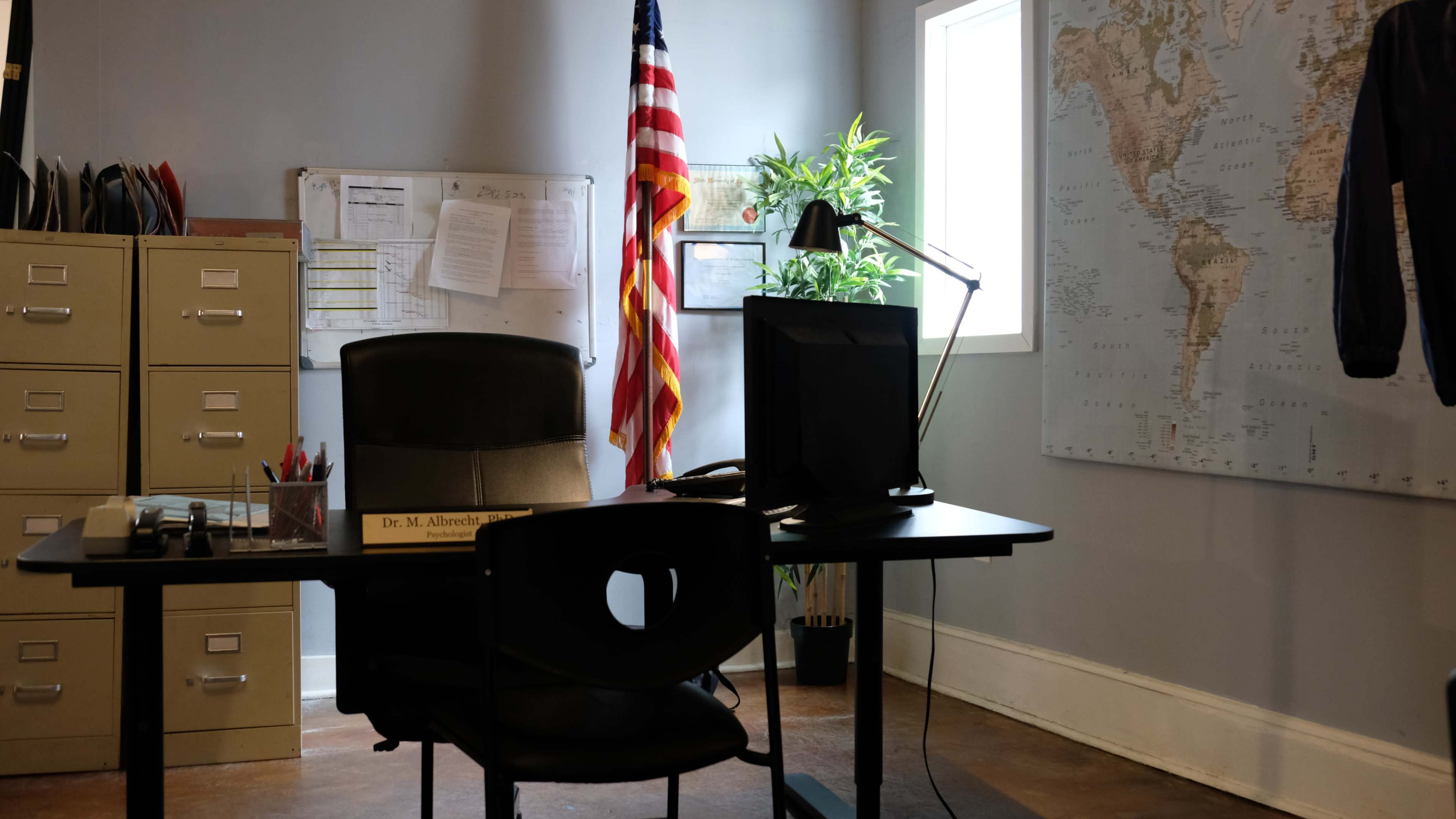 The image shows an office with a desk, computer, chair, American flag, and a map on the wall.