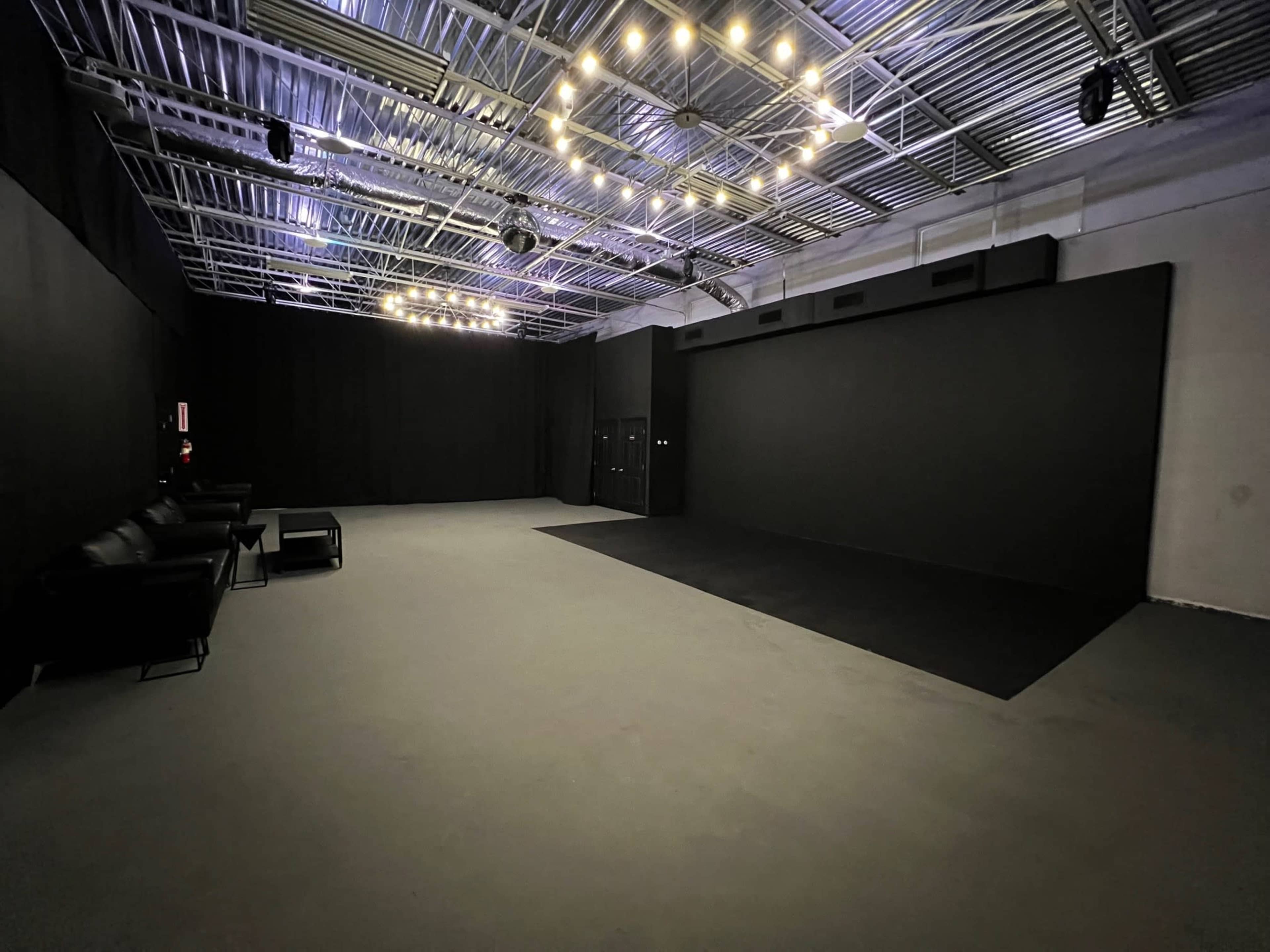 An empty studio space with black walls, a dark floor, and overhead lights creating an illumination effect.