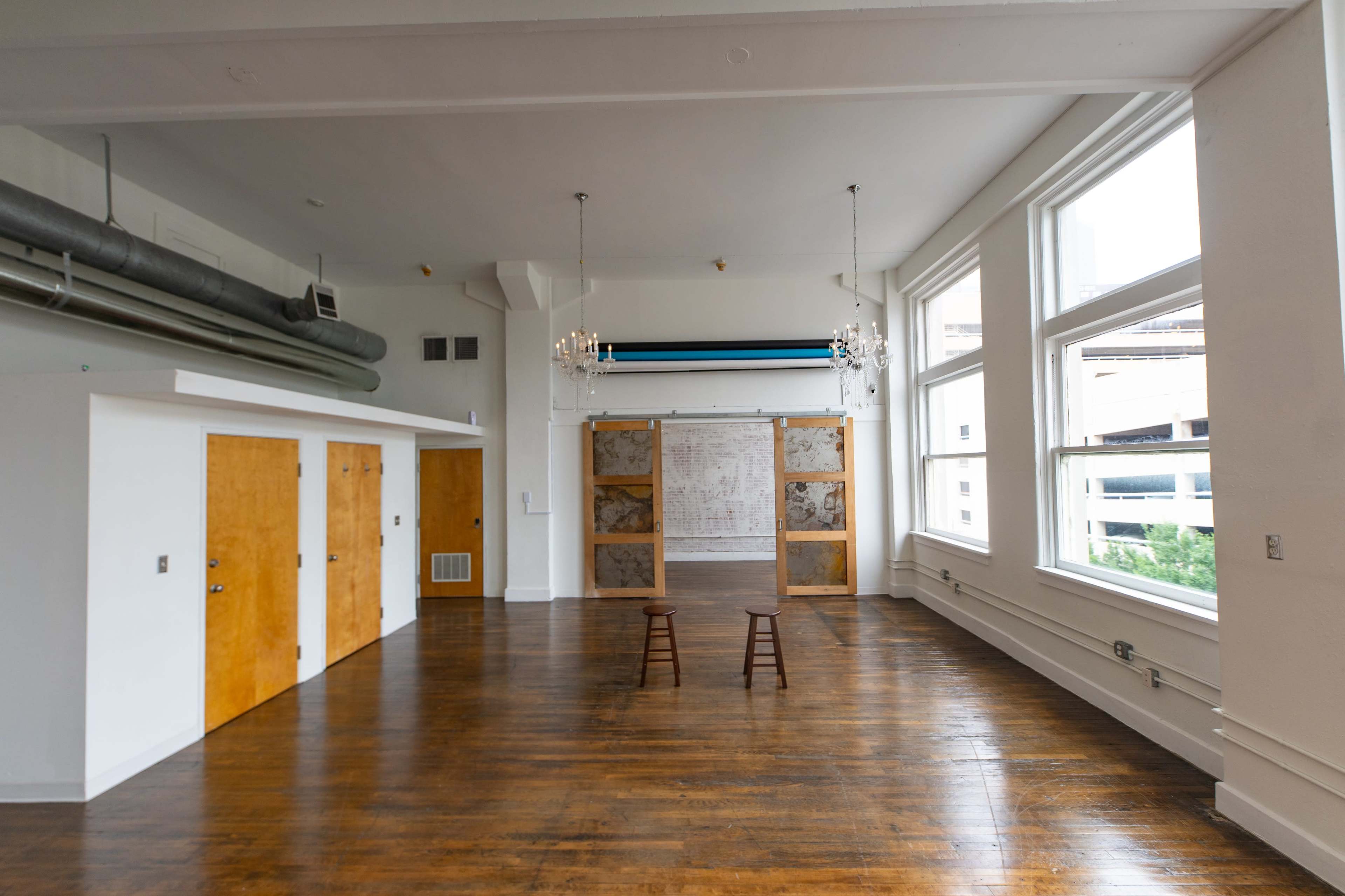 The image shows a spacious, well-lit room with wooden flooring, two stools facing a divider, and large windows allowing natural light to enter.