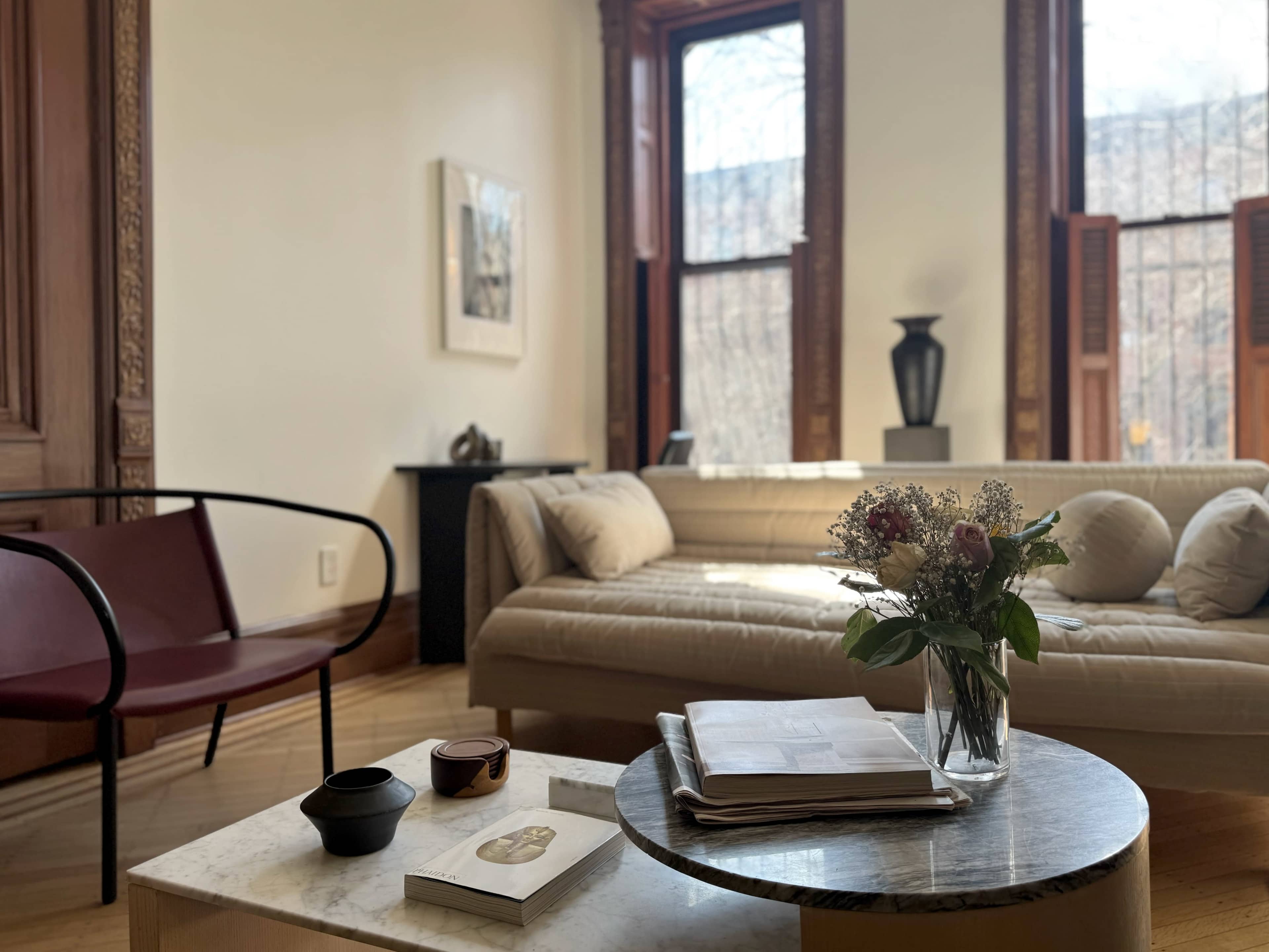 The image shows a well-lit living room with a beige sofa, a round marble coffee table adorned with magazines and a vase of flowers, and large windows with wooden frames.