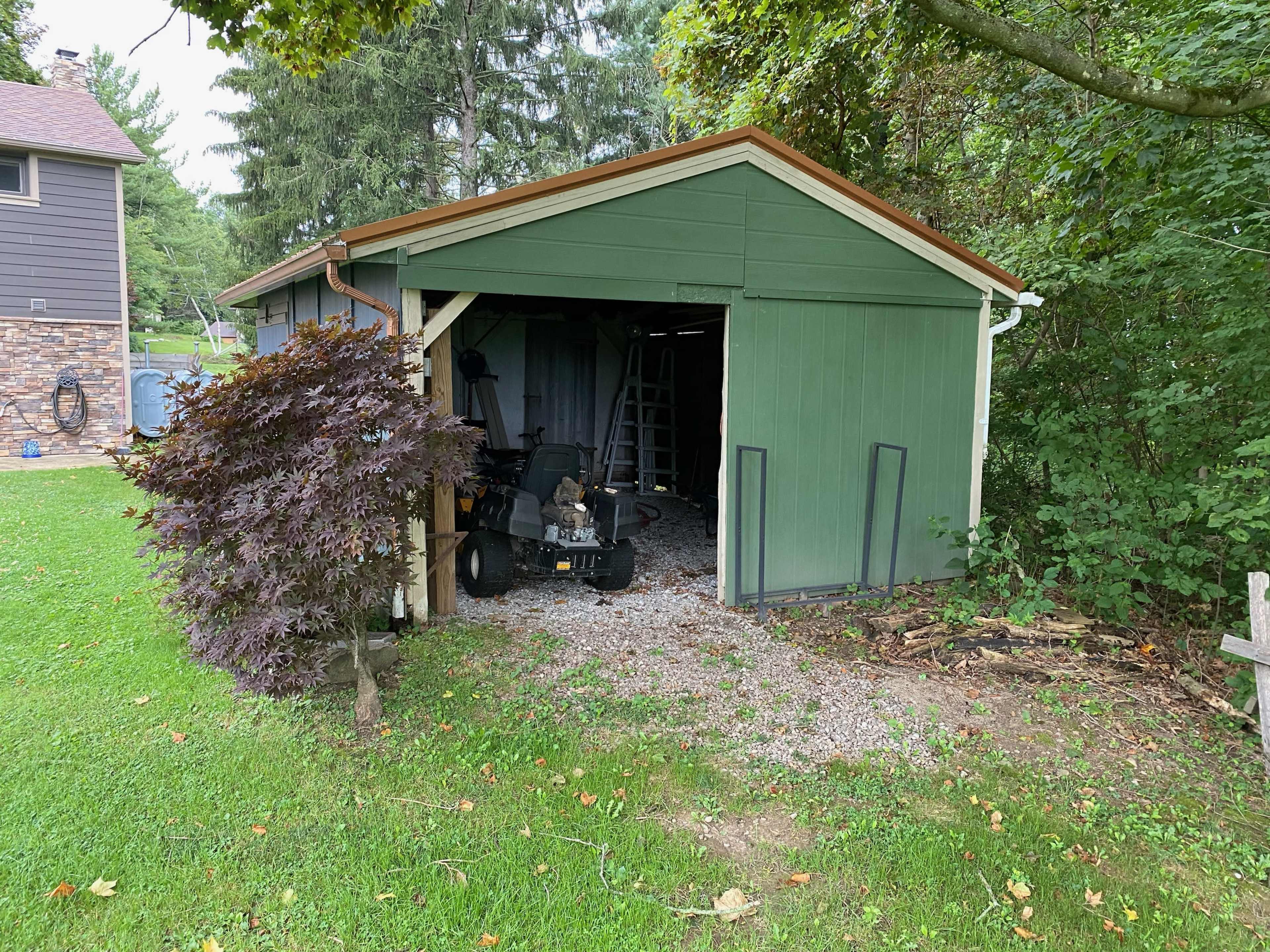 A green shed with an open doorway sits on a grassy area surrounded by trees and a small shrub.