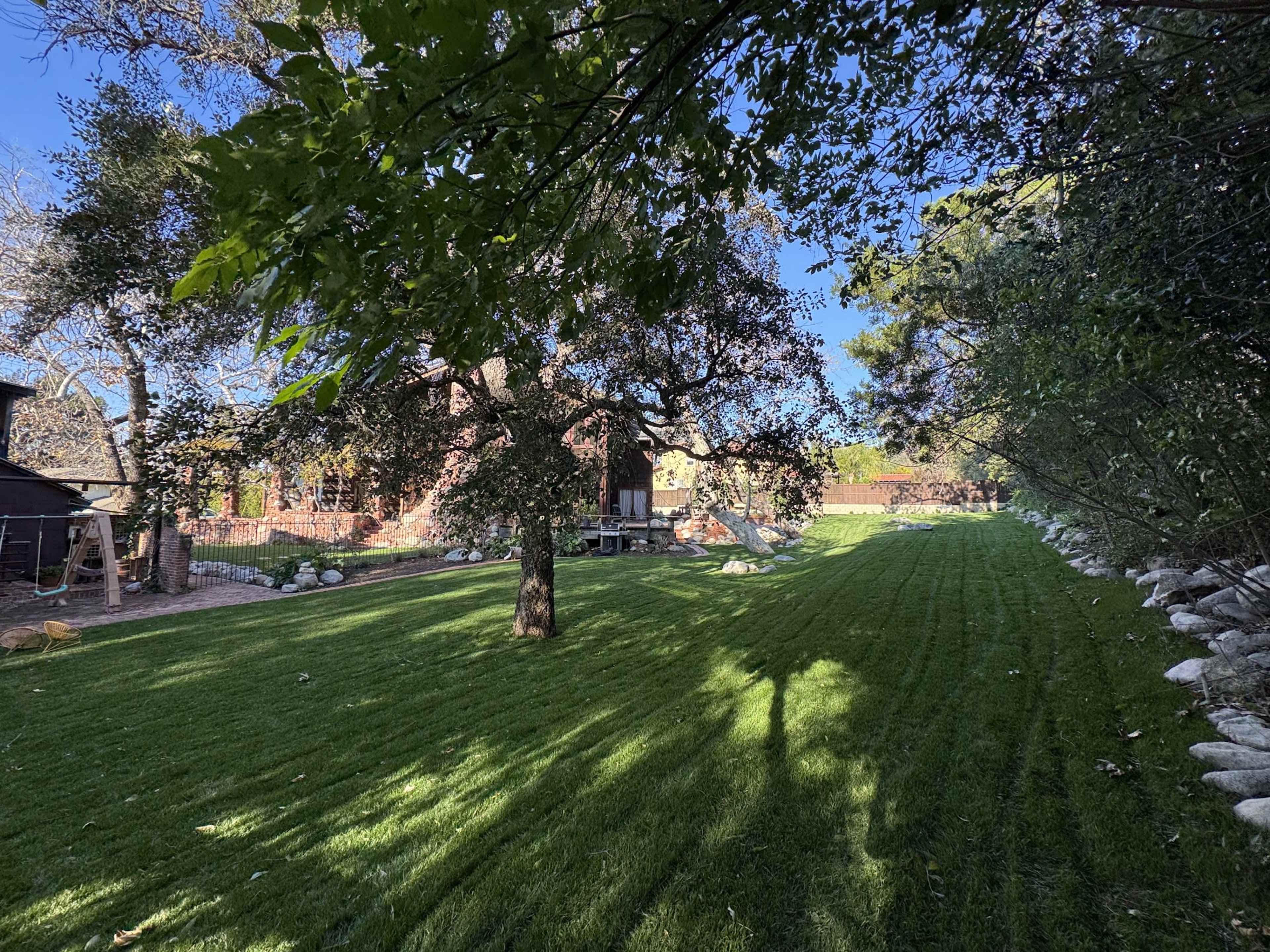 The image shows a neatly manicured lawn with a tree in the foreground and a building in the background, surrounded by rocks and greenery.