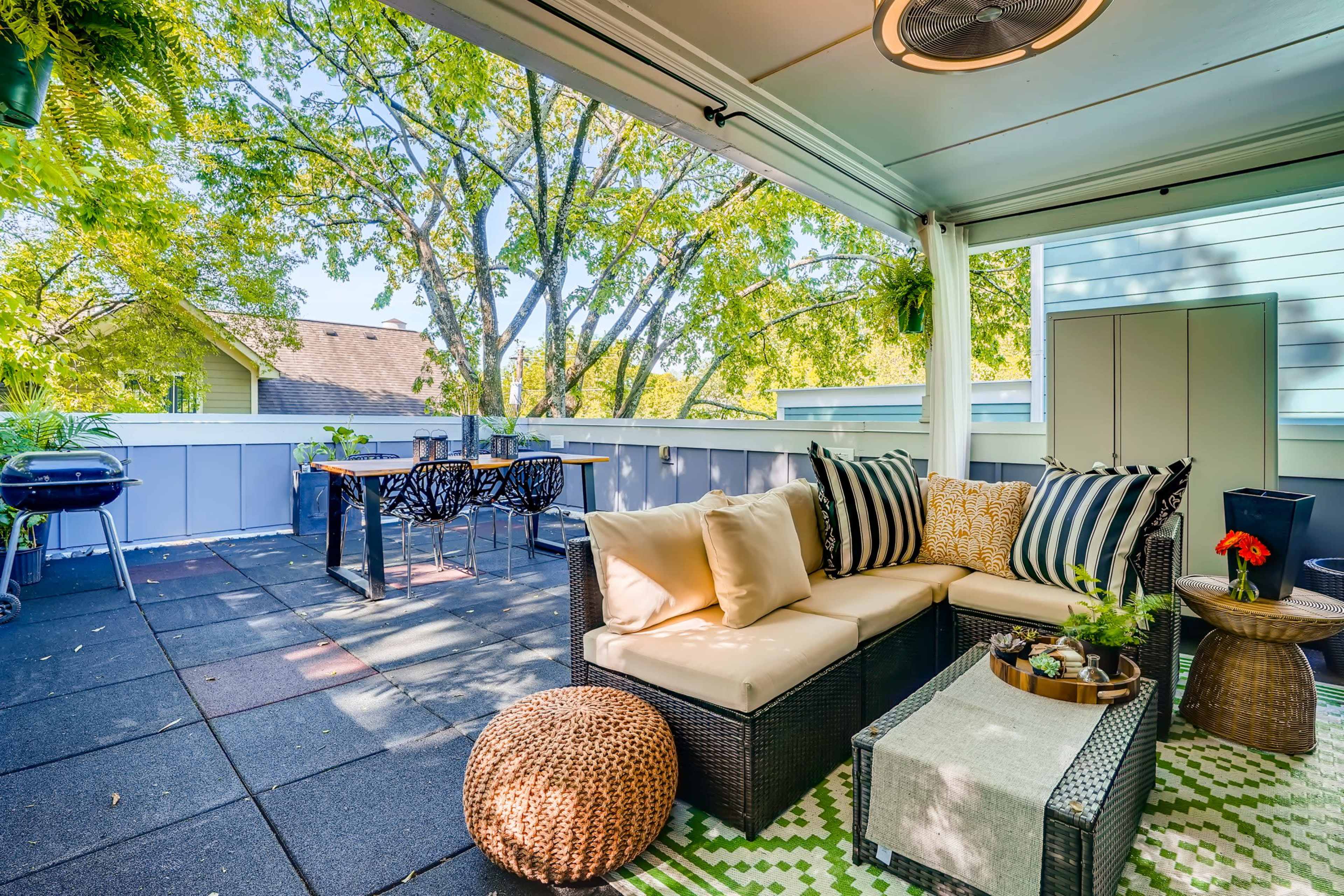 A cozy outdoor patio features a sectional sofa, a coffee table with decorative items, a dining area, and a grill, all surrounded by greenery.
