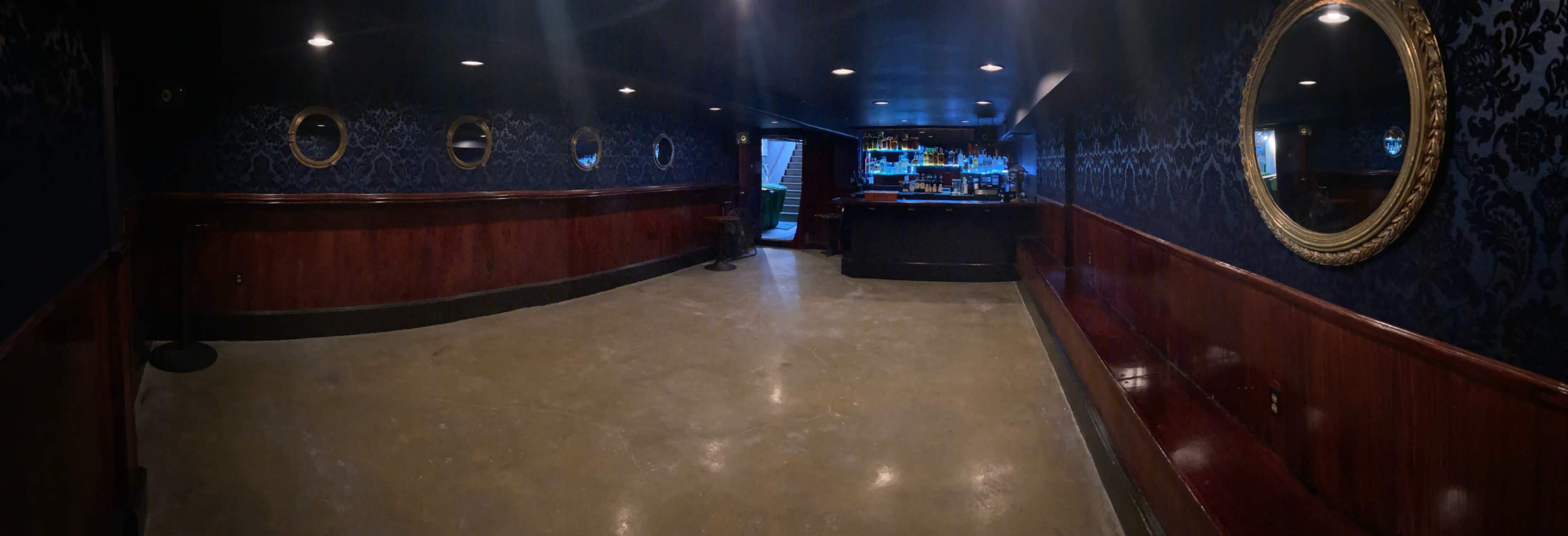 A dimly lit bar interior with dark wood paneling, ornate mirrors, and a visible bar counter at the back.
