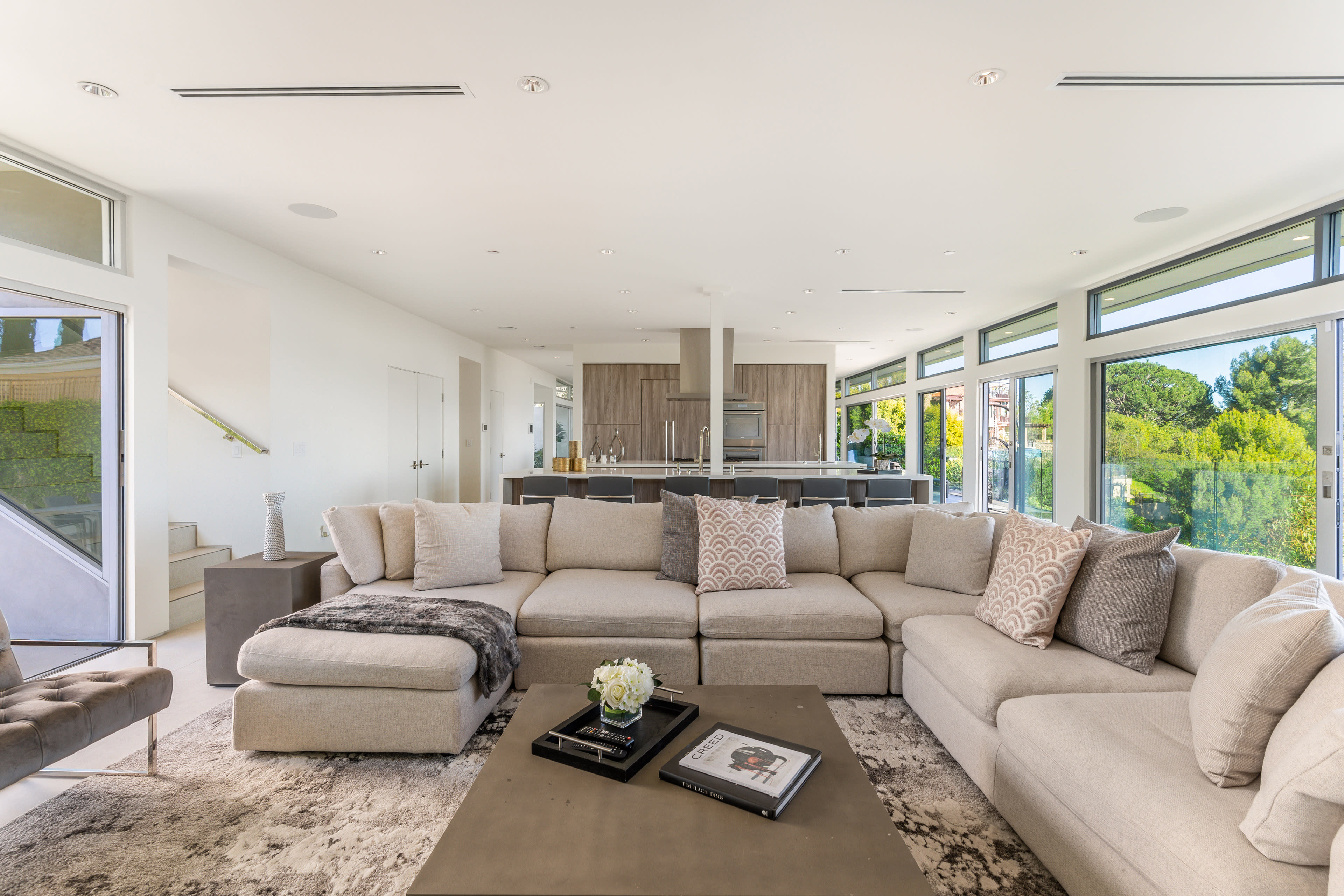 The image shows a spacious, modern living room with a large sectional sofa, a coffee table, and large windows that overlook a garden.