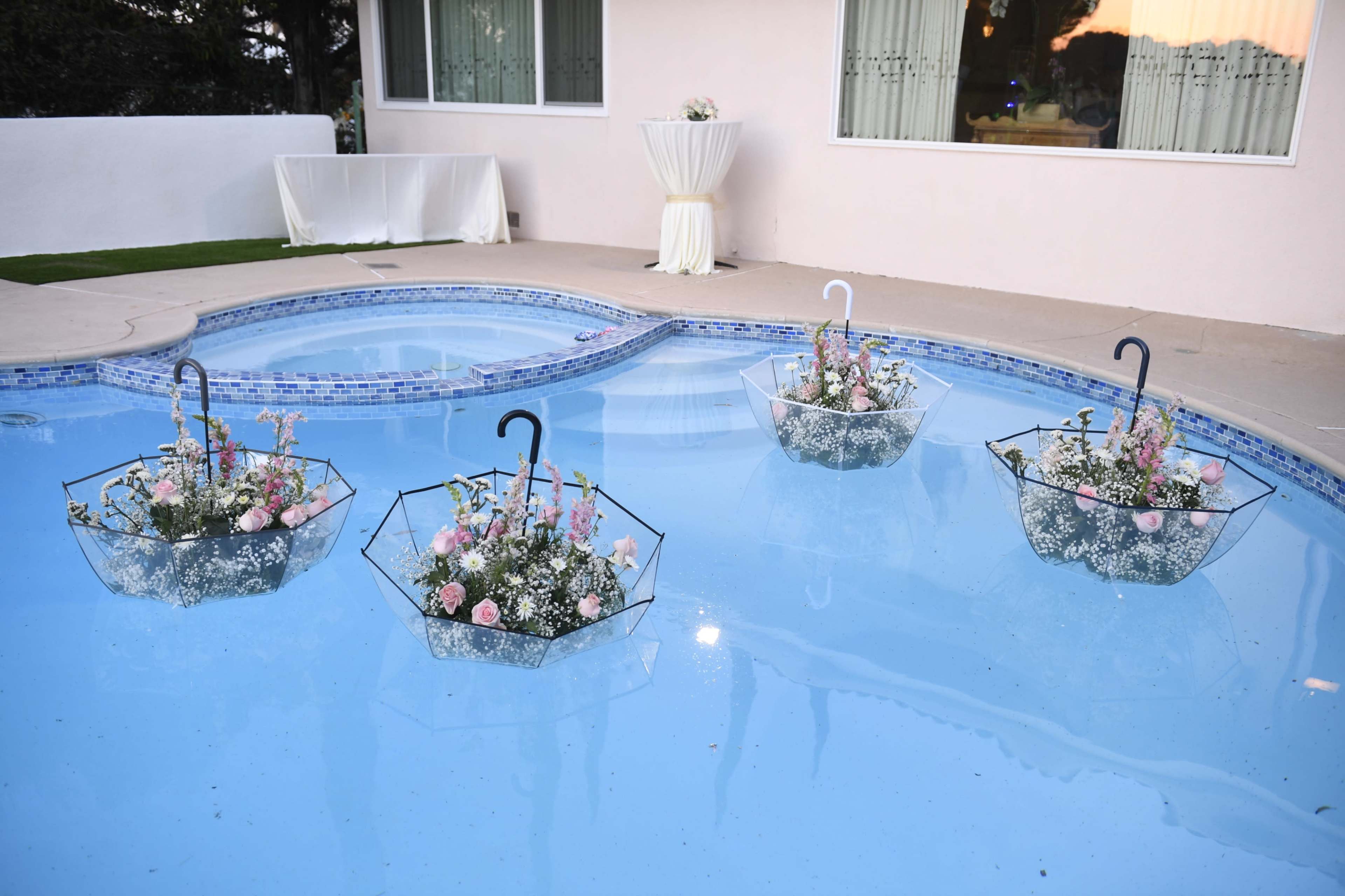 Four transparent umbrellas filled with flowers float in a swimming pool next to a white wall and a table set up for an event.