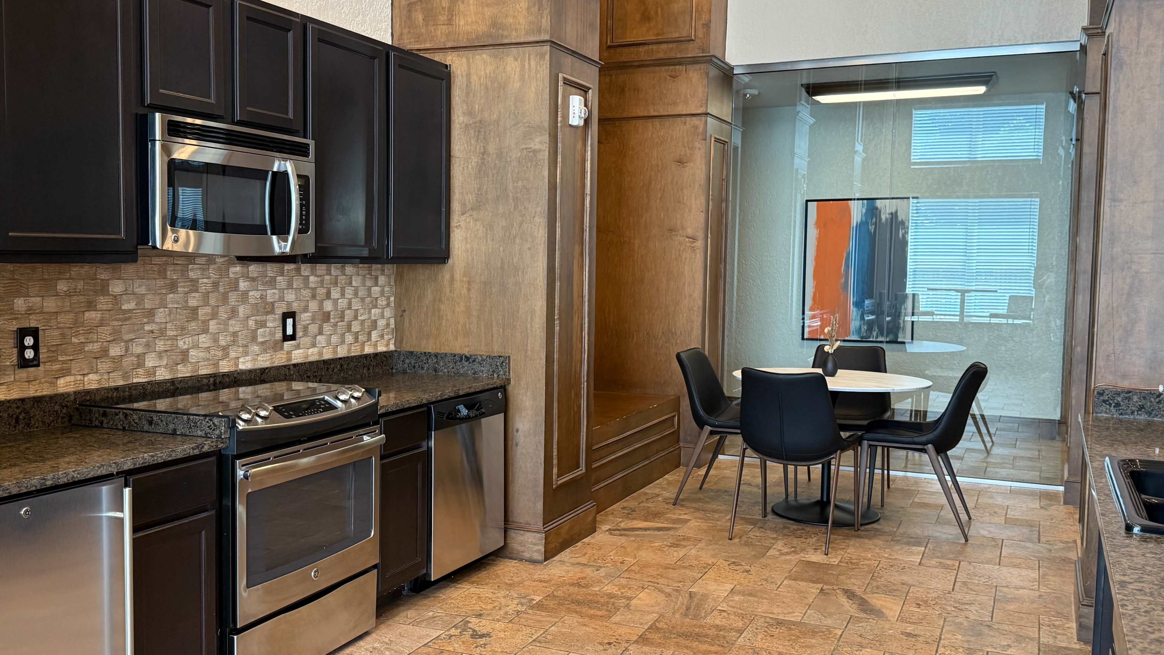 The image shows a modern kitchen with dark cabinetry, stainless steel appliances, and a dining area featuring a round table and black chairs adjacent to a glass-walled room.