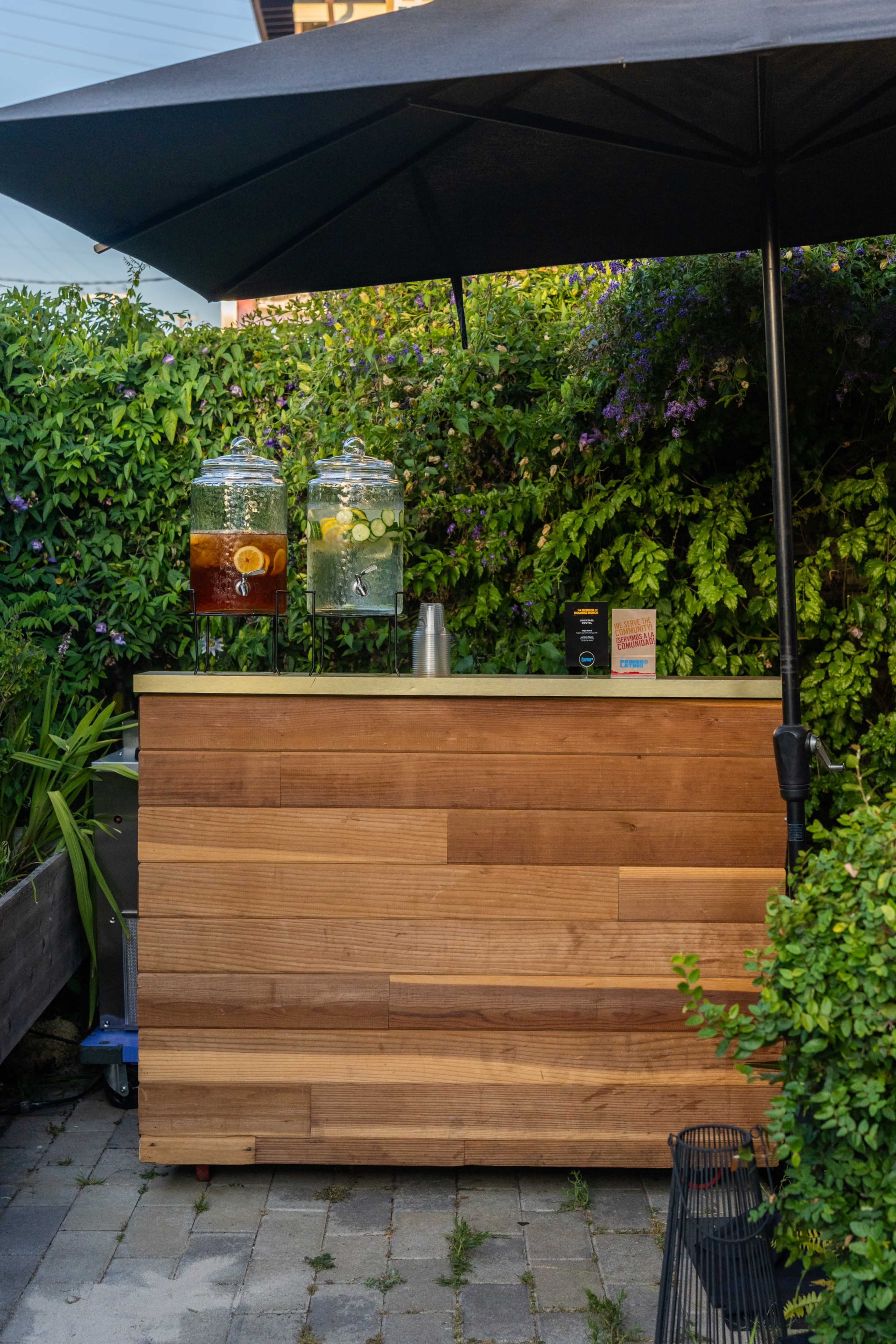 A wooden drink stand with two glass dispensers filled with beverages is set up under a black umbrella surrounded by lush greenery.