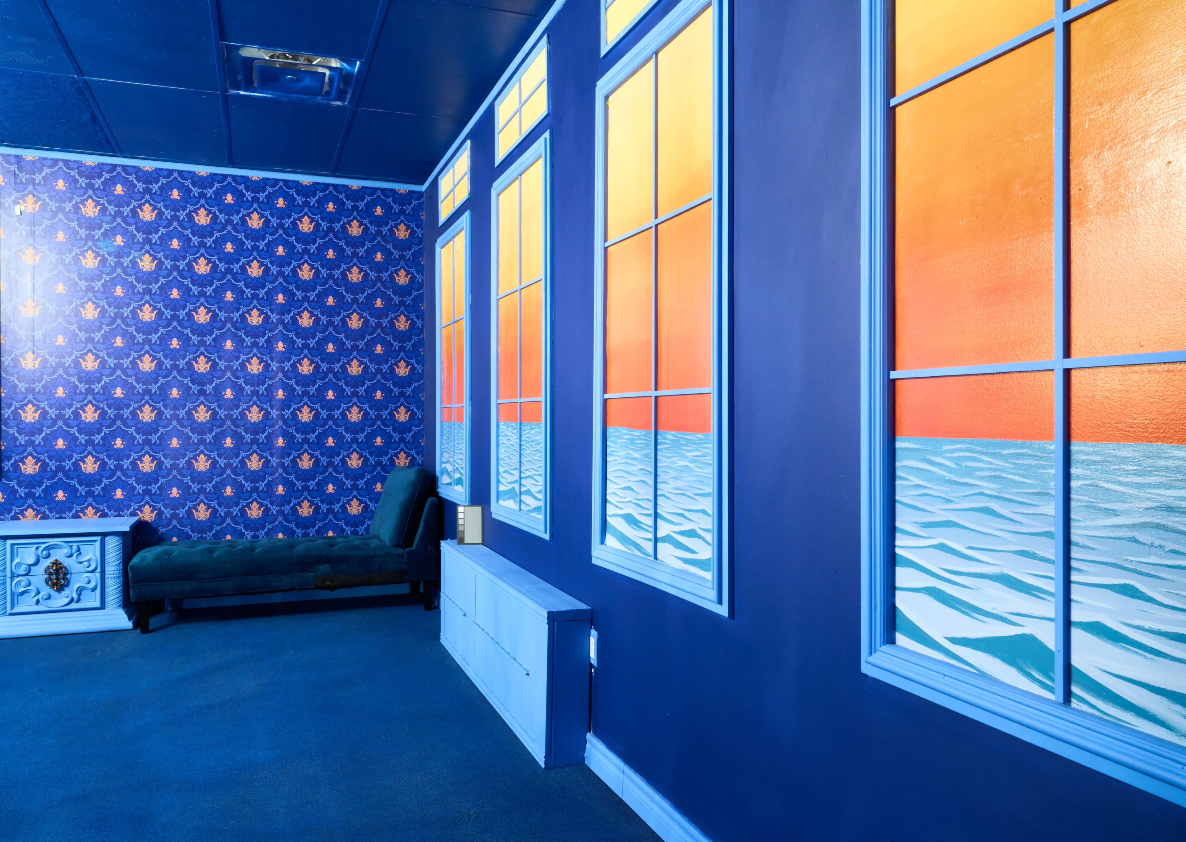 The image shows a room with blue walls adorned with a patterned wallpaper, featuring large window murals that depict a sunset over water, alongside a green sofa and a white storage unit.