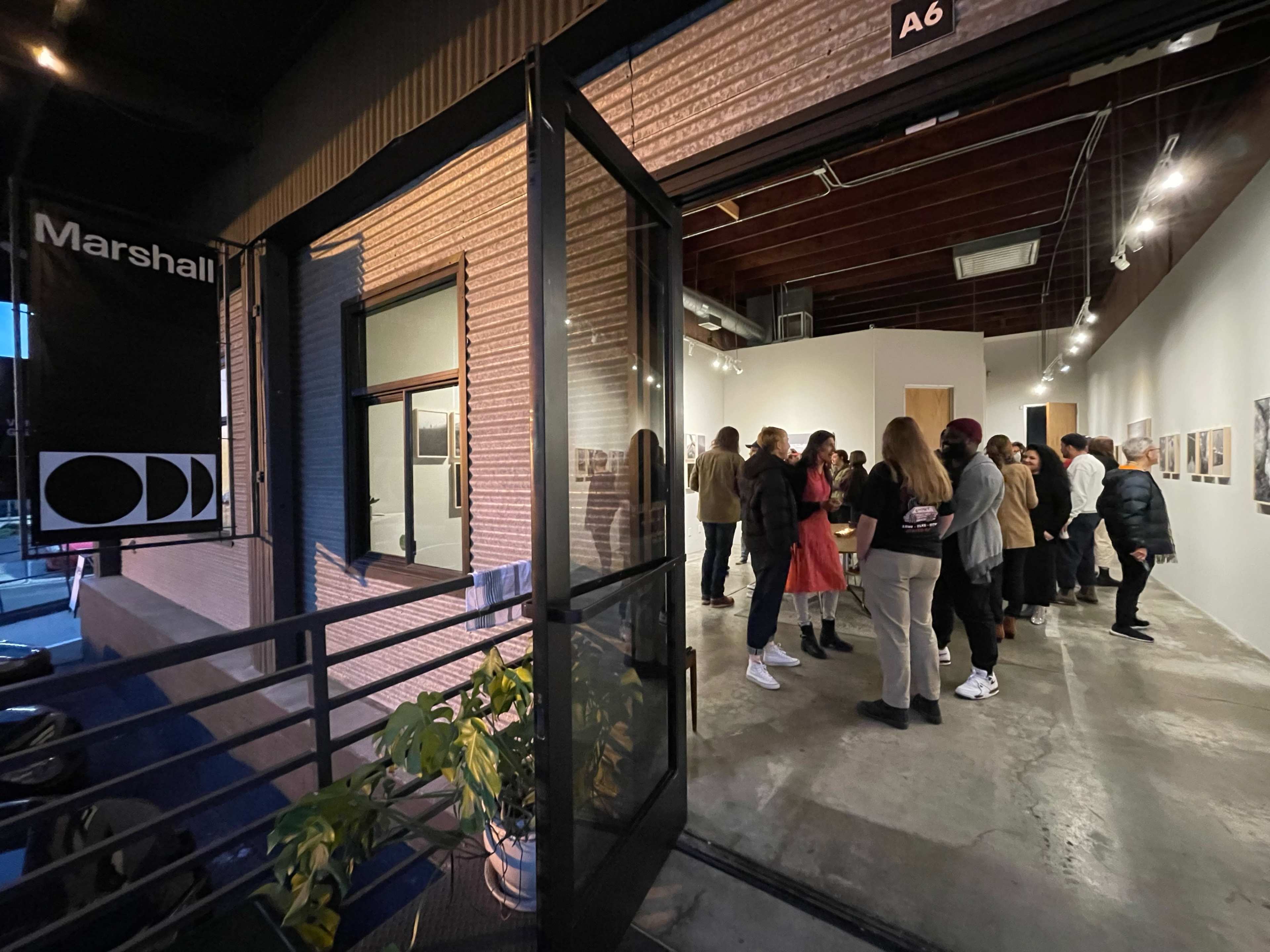 A group of people gathers inside an art gallery, engaged in conversation, while a doorway opens to the outside.