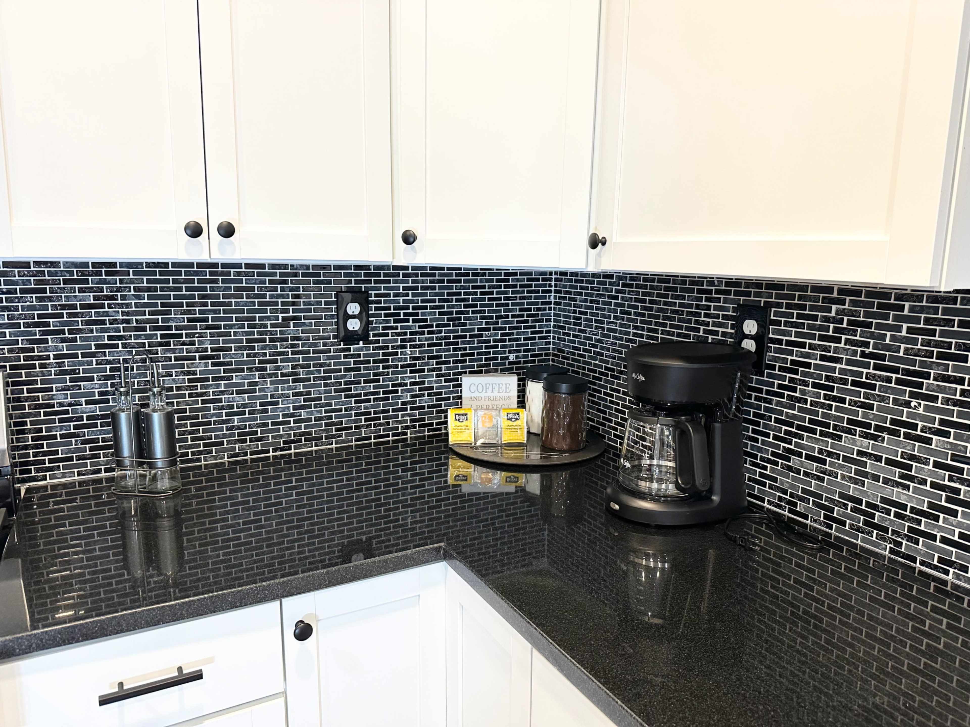 A modern kitchen countertop features black tile backsplash, a coffee maker, and various kitchen items arranged neatly.
