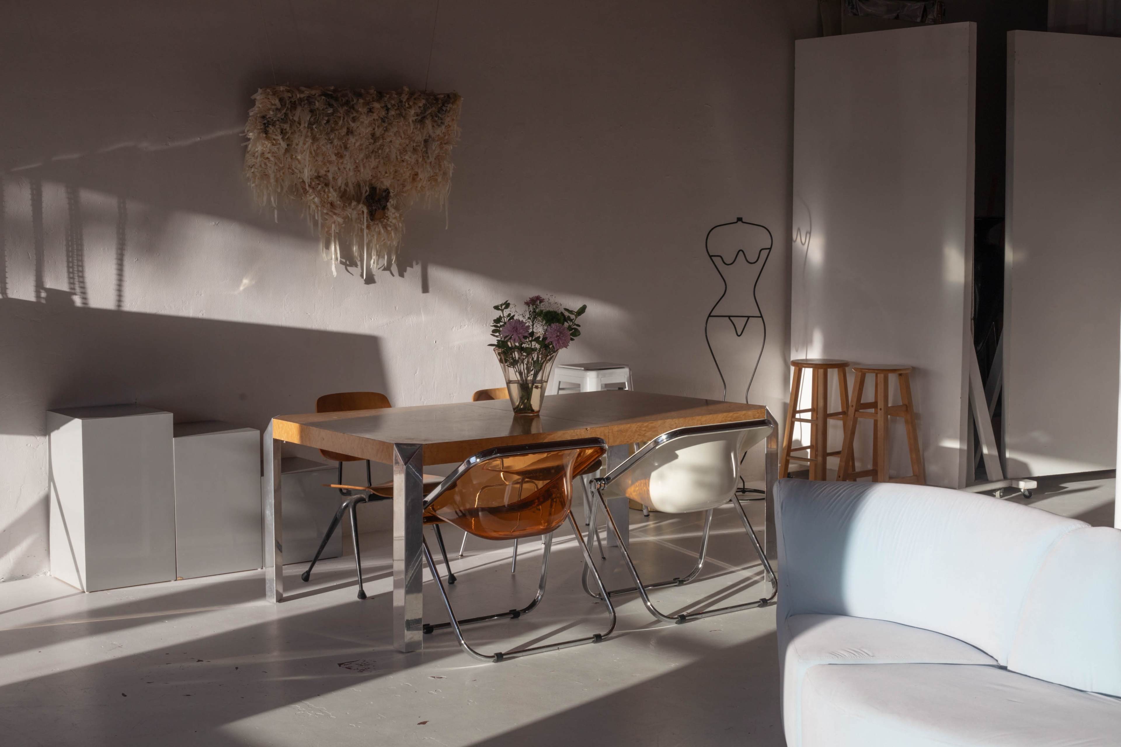 A minimalist interior features a wooden dining table surrounded by chairs, with a vase of flowers on the table and a minimalist sculpture in the background.