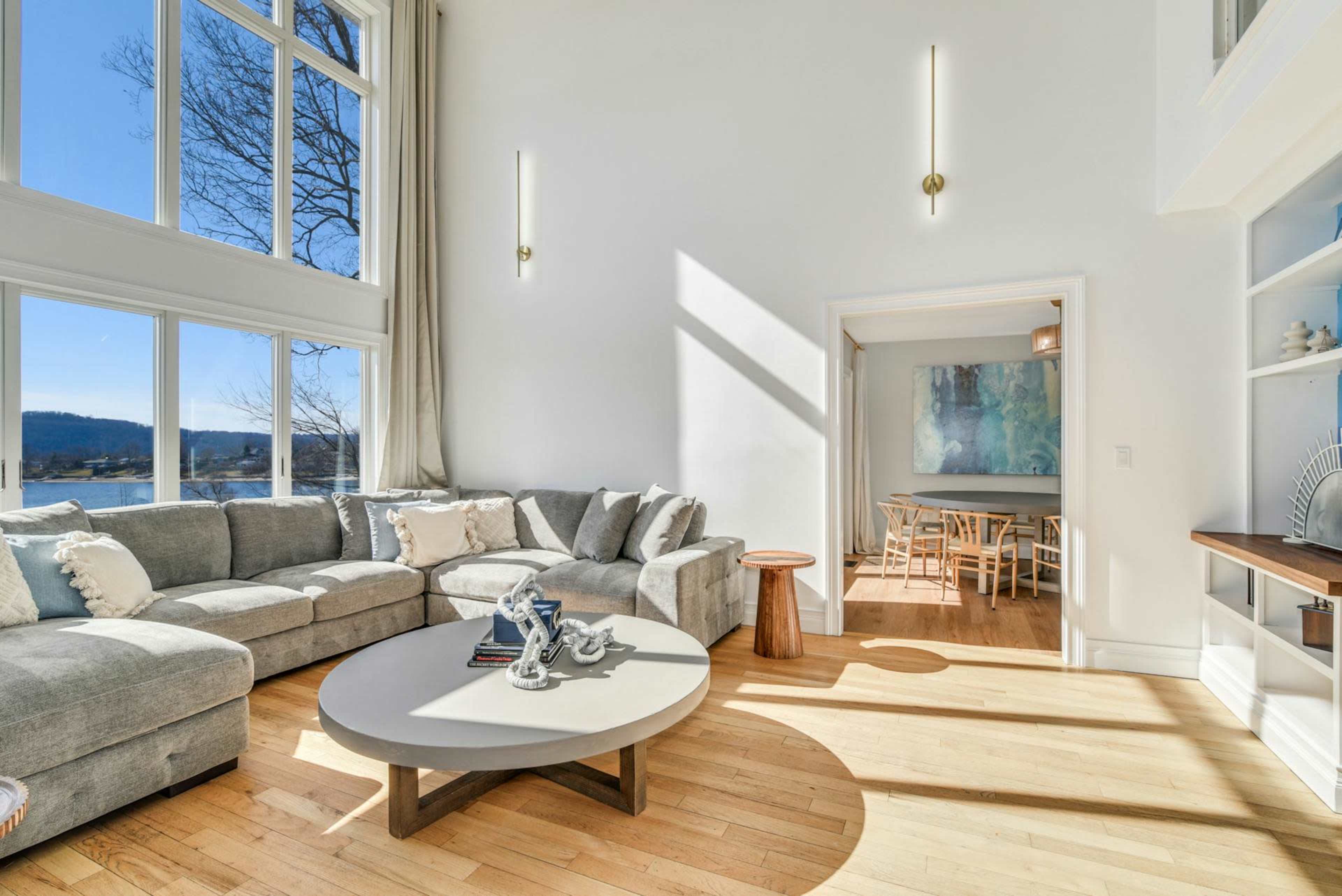 A bright living room features a large sectional sofa, a circular coffee table, and a dining area visible through an open doorway.