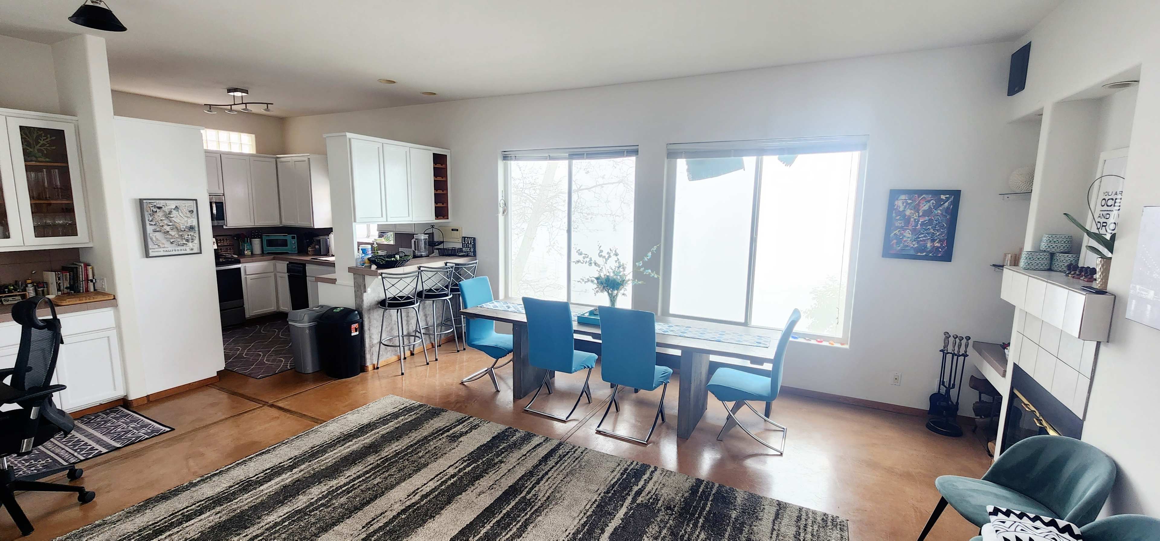 The image shows a bright, open-concept living area featuring a dining table with blue chairs, a kitchen area, and large windows that let in natural light.