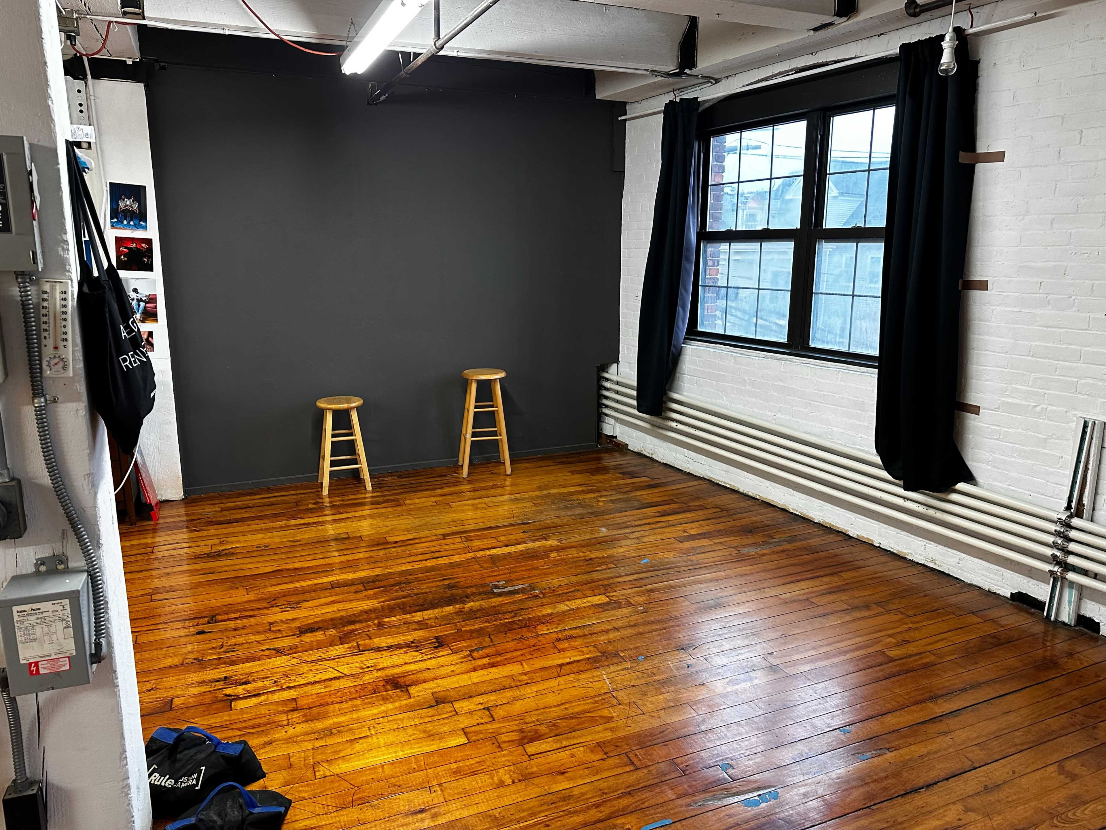 A minimalist room with wooden floors, two wooden stools, a gray wall, and a large window with black curtains.