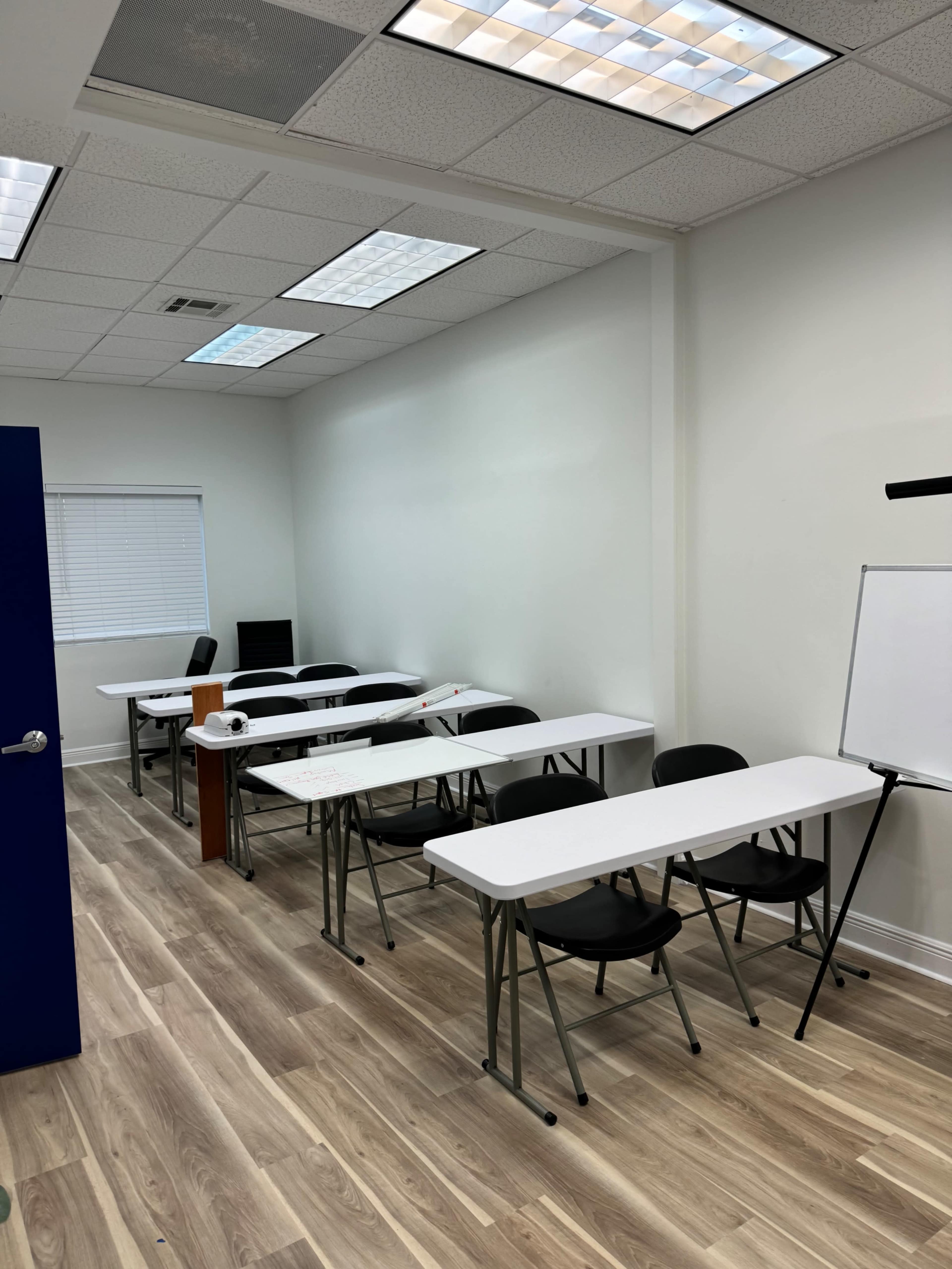 An empty classroom with several rows of tables and chairs, a whiteboard, and a window allowing natural light.
