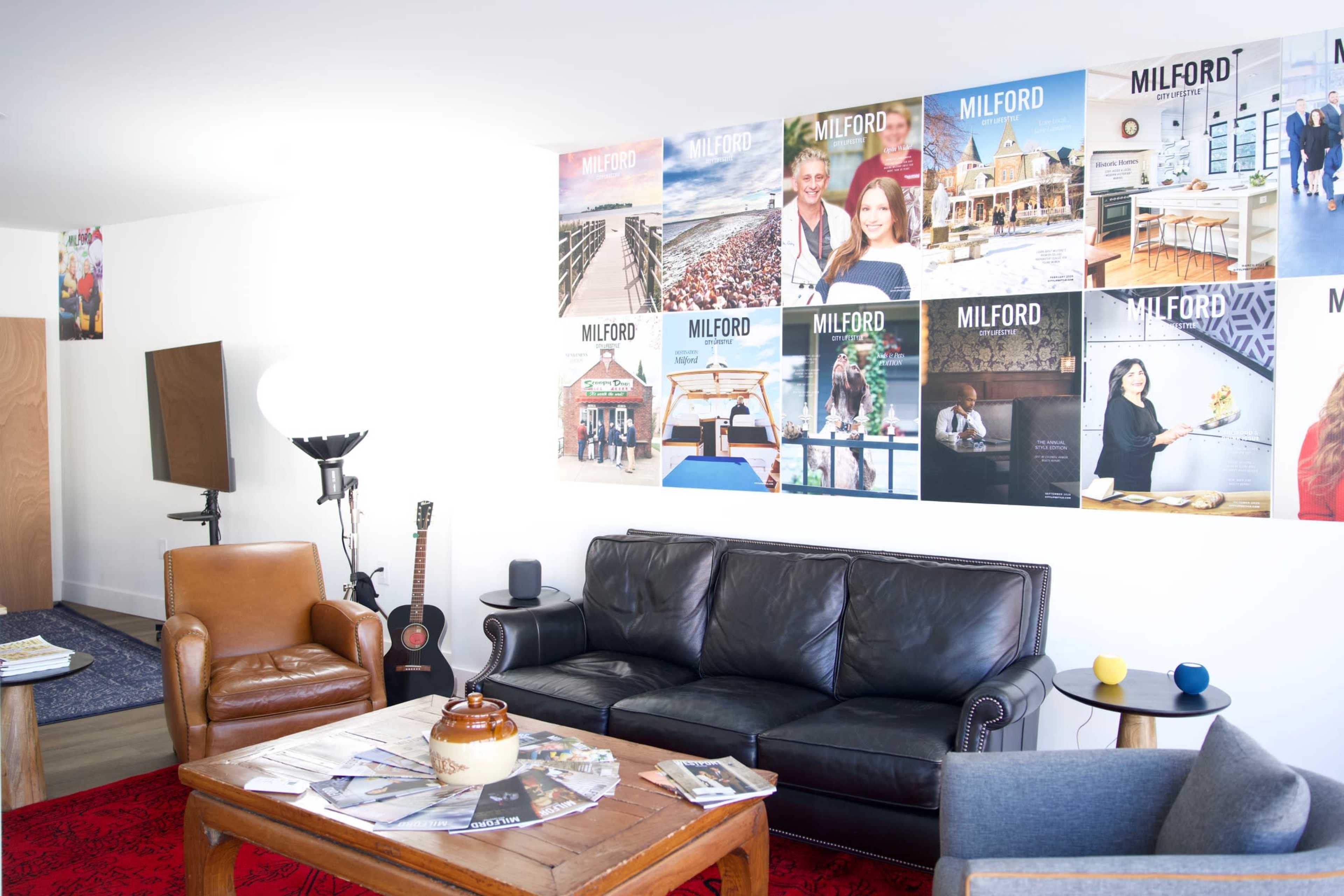 The image shows a modern living space featuring a dark leather sofa, a wooden coffee table with a decorative item, and a wall adorned with various photographs and posters.