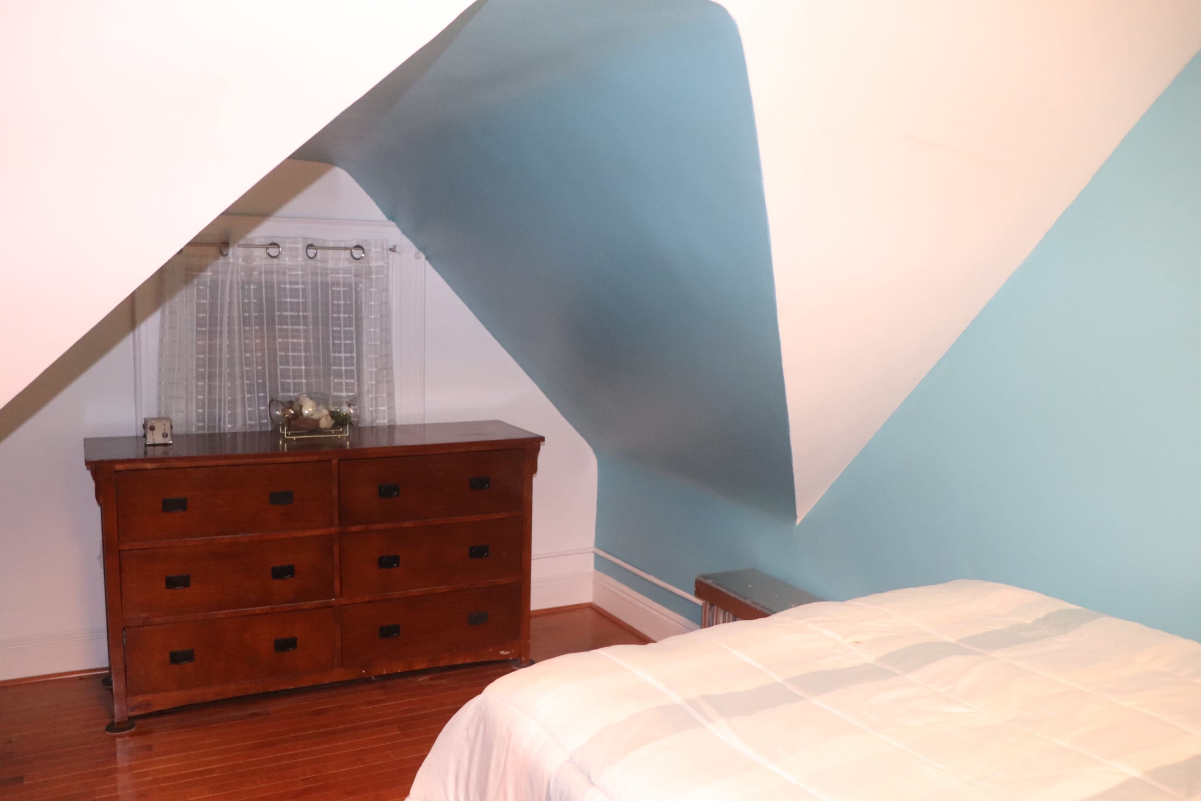 A bedroom with a slanted ceiling features a wooden dresser and a bed with a white coverlet.