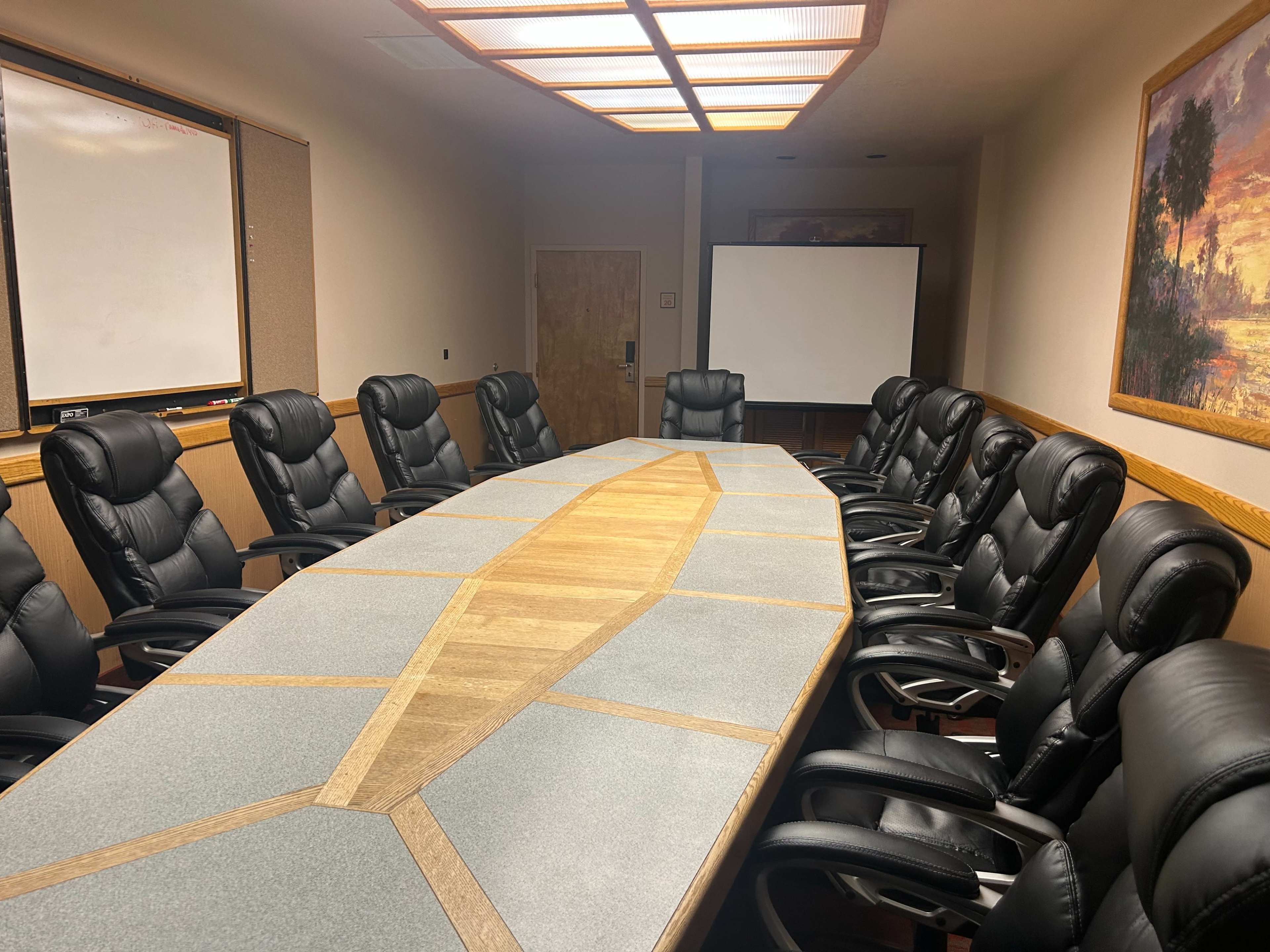 The image shows a conference room featuring a large oval table surrounded by black leather chairs, with a projector screen and whiteboard in the background.