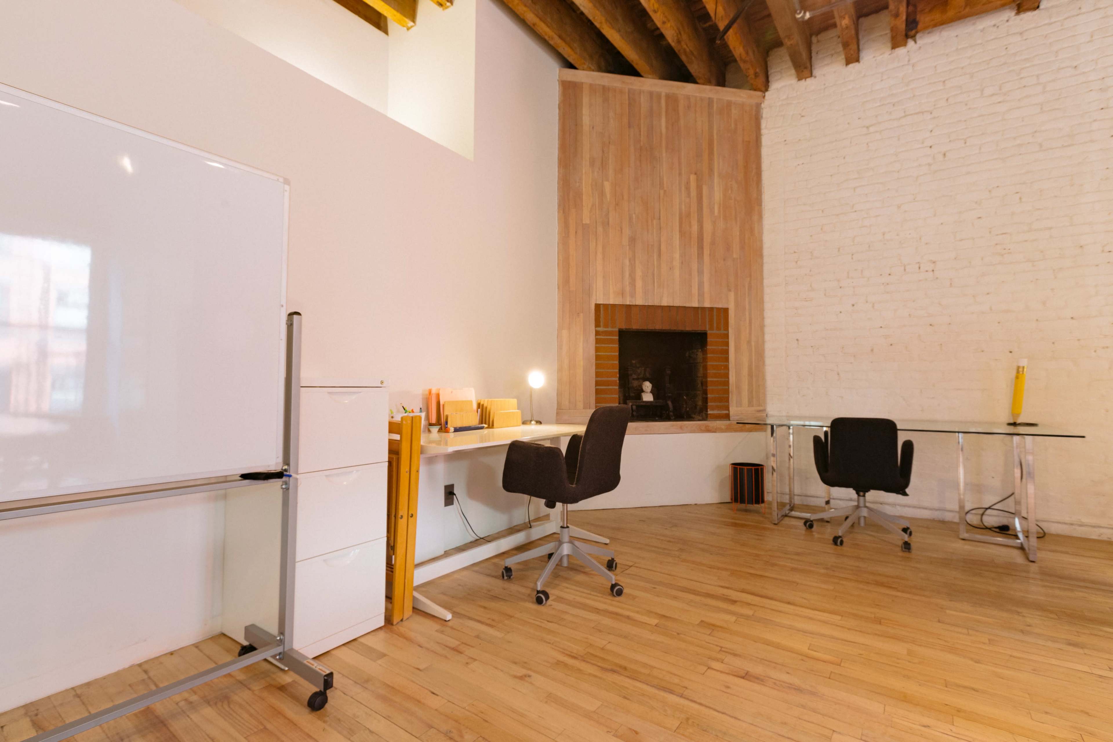 A modern office space features two desks and a whiteboard, with exposed wooden beams and a brick wall.