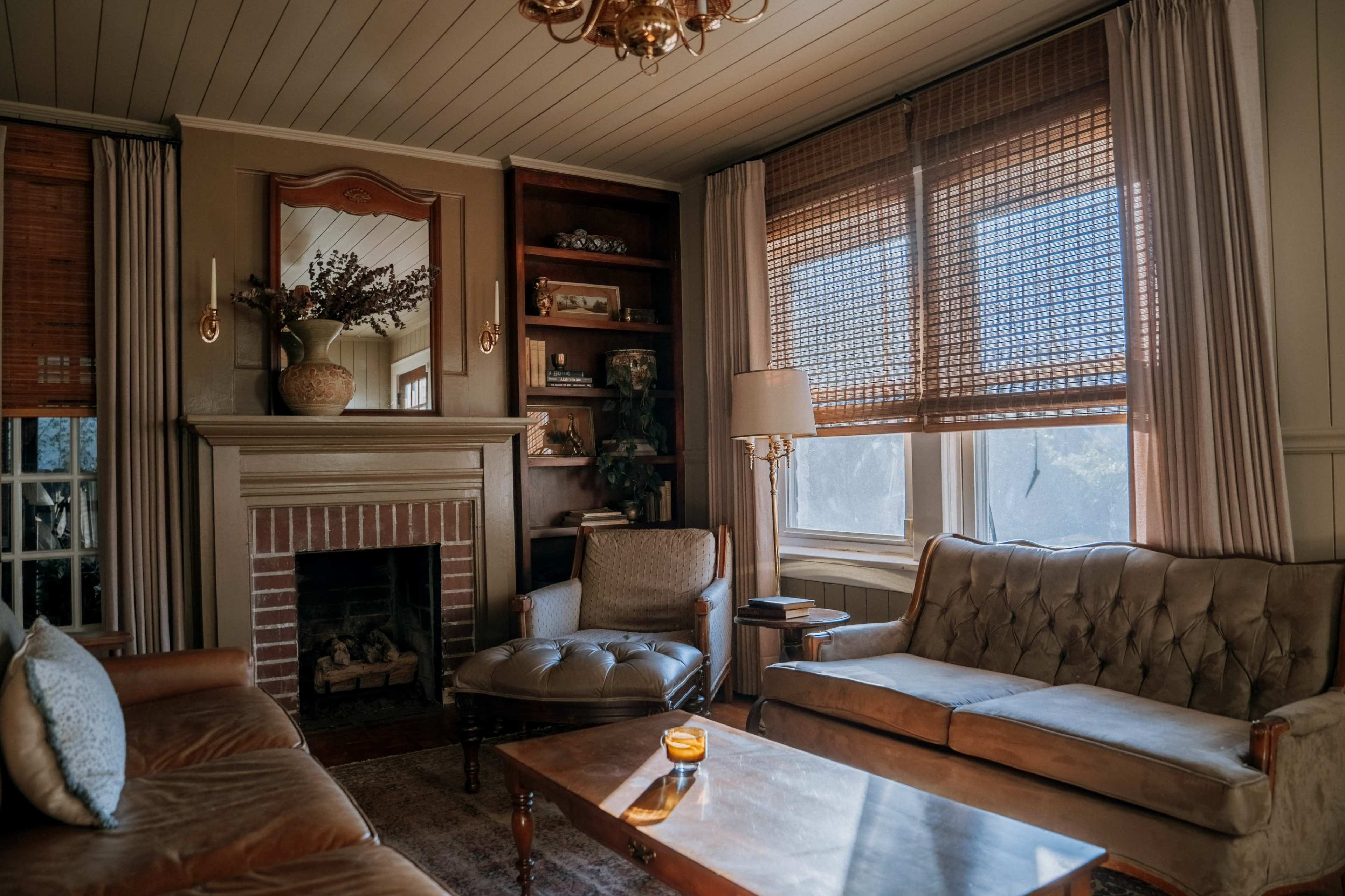 The living room features a fireplace, a brown sofa, an armchair, and a wooden coffee table, with large windows covered by wooden blinds allowing natural light to enter.