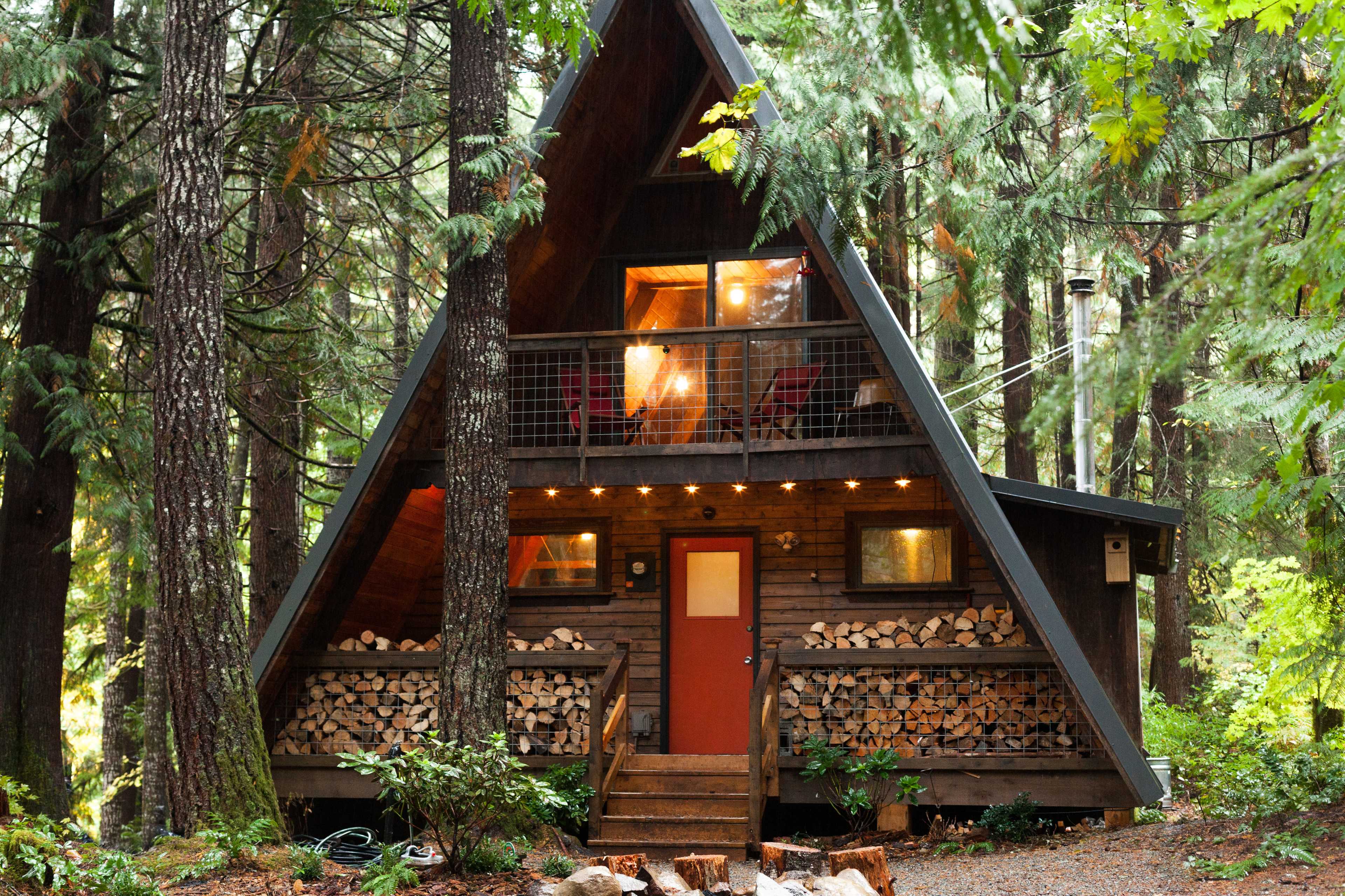 A wooden A-frame cabin with a red door is surrounded by tall trees and features stacked firewood on its front porch.