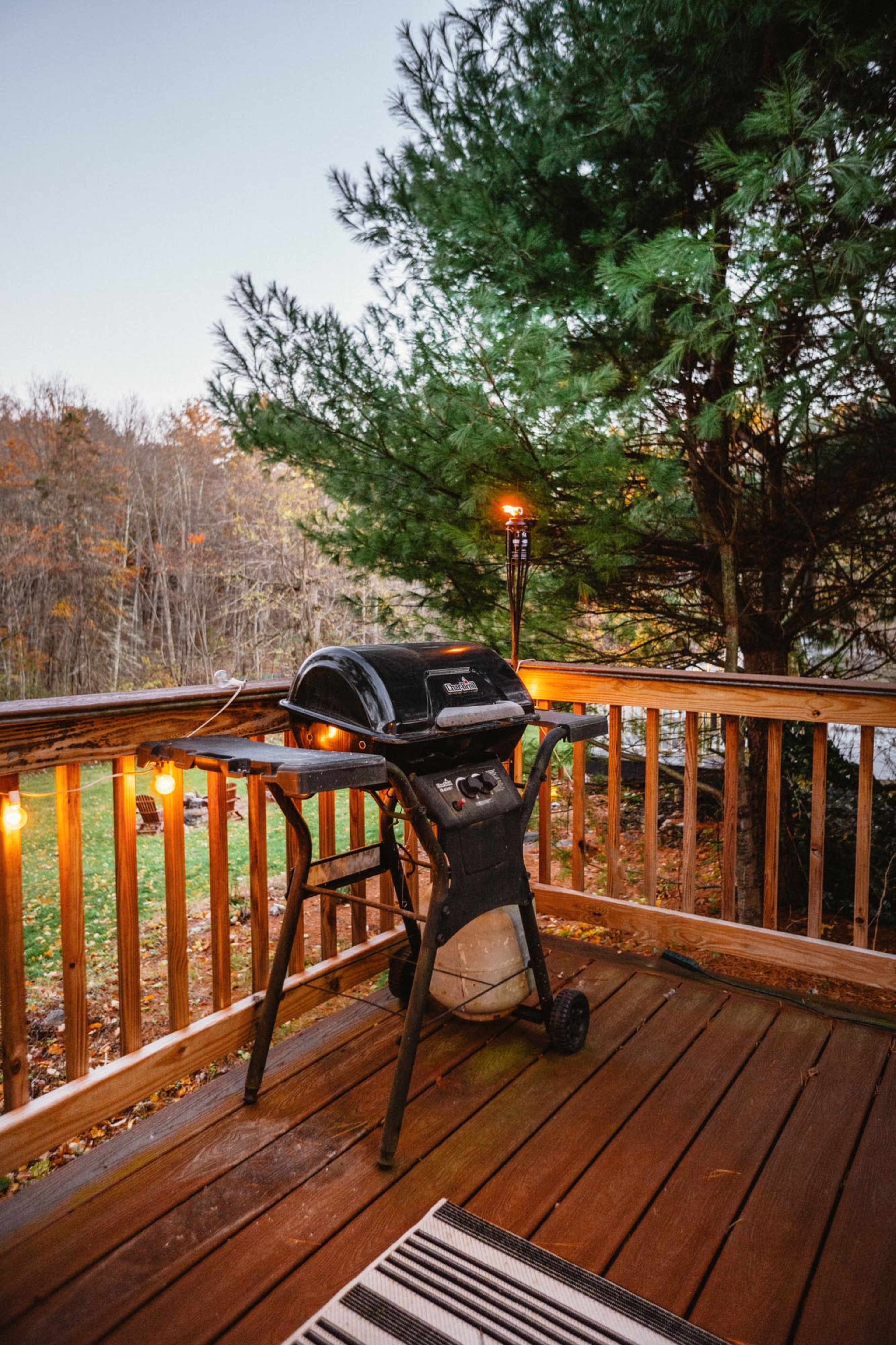 A gas grill is set up on a wooden deck surrounded by trees, with string lights illuminating the area at dusk.